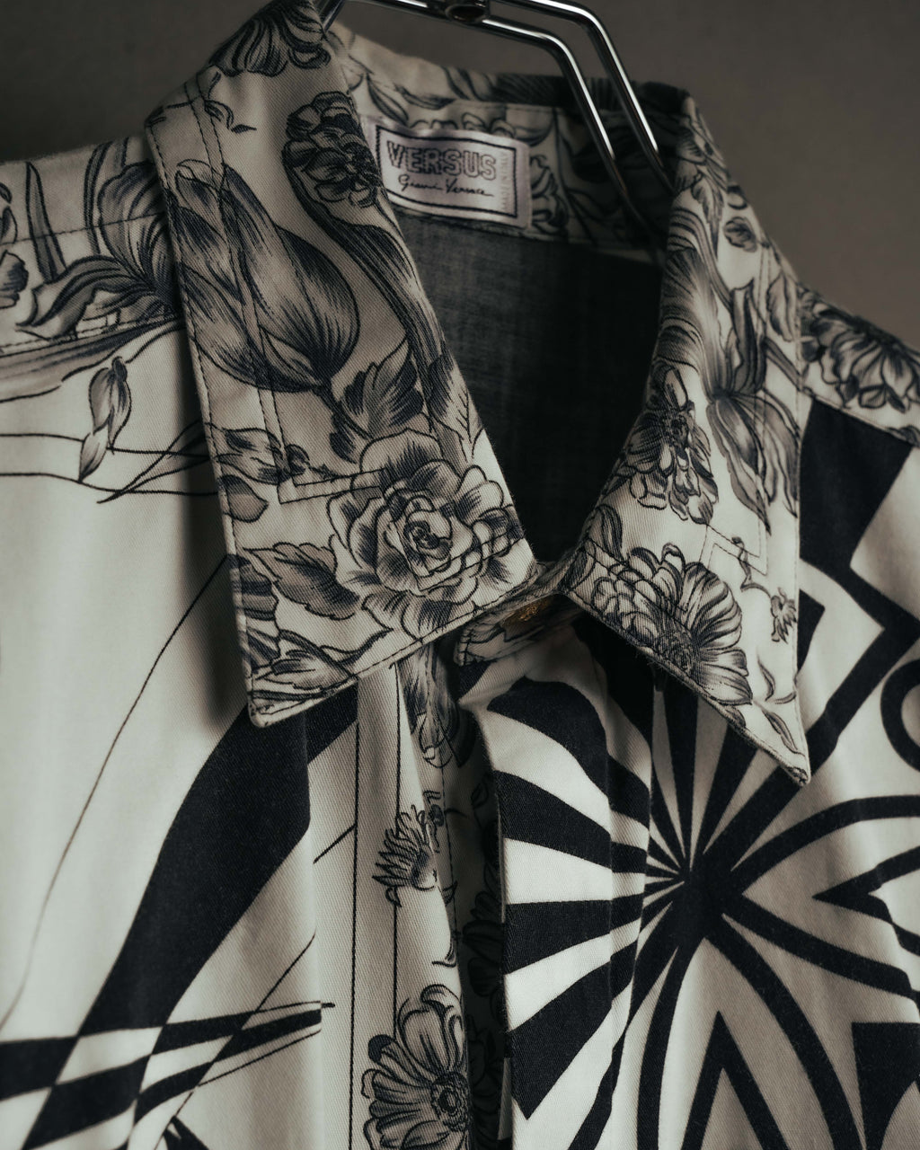 白系の織り地に黒い二重枠を配したVERSUS Gianni Versaceの旧形式ラベルと、線描の花柄を配した襟元を捉えたカット。身頃にはモノトーンのオプティカルパターンが続く。