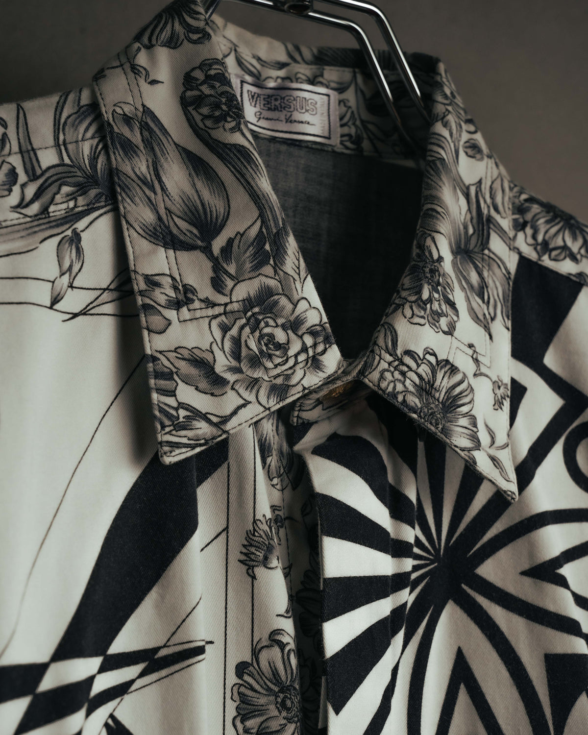 白系の織り地に黒い二重枠を配したVERSUS Gianni Versaceの旧形式ラベルと、線描の花柄を配した襟元を捉えたカット。身頃にはモノトーンのオプティカルパターンが続く。