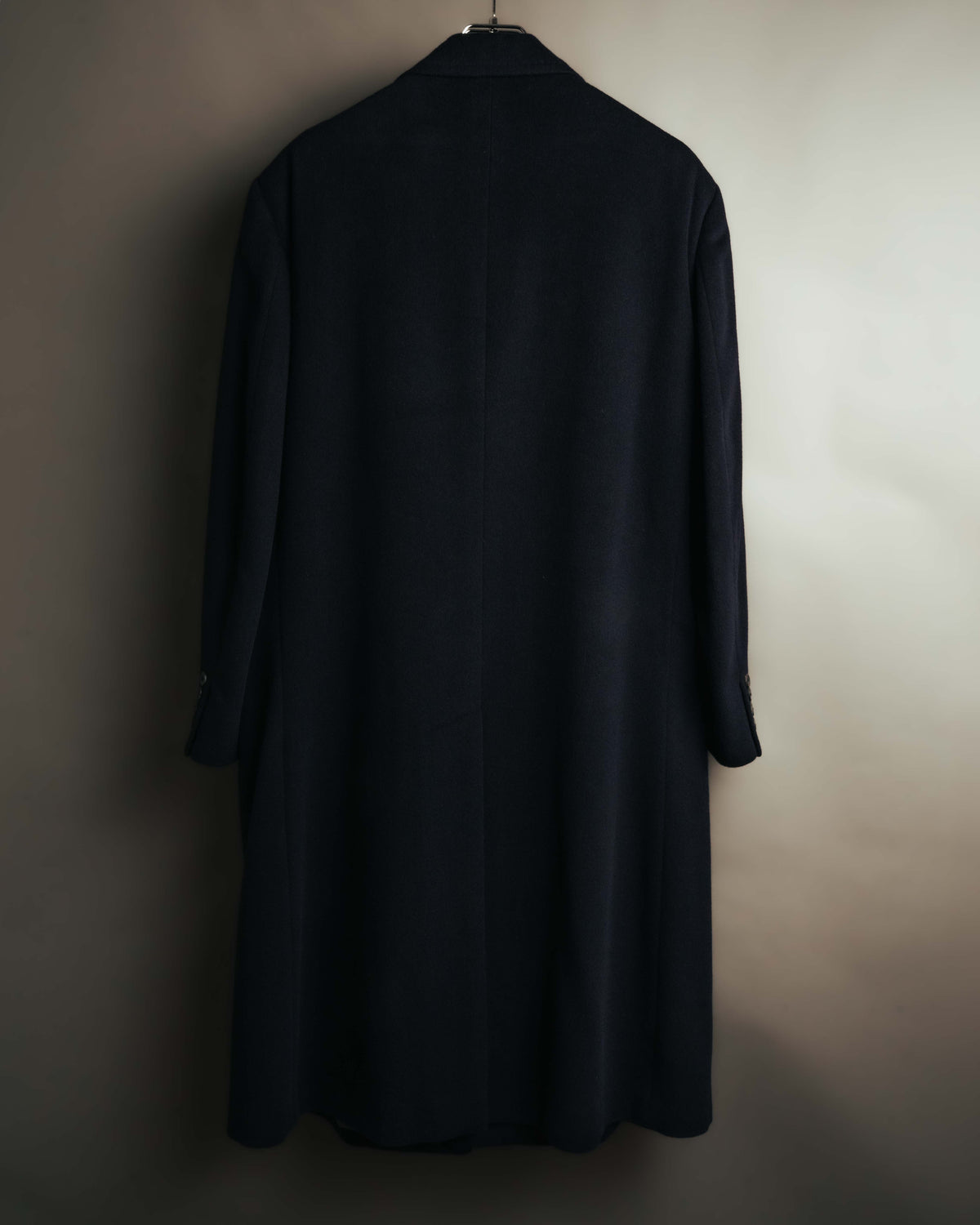 黒に近いダークネイビー系のロングコートを背面から捉えた全体カット。縦へまっすぐ落ちるラインと、裾に入ったセンターベントを確認できます。