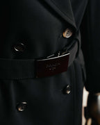 共布ベルトに配された、PRADA MILANO DAL 1913の文字と紋章が刻まれた黒系メタルの大型バックルを捉えたディテールカット。周囲にはダブルブレストのボタンとフラップポケットの縁が写ります。