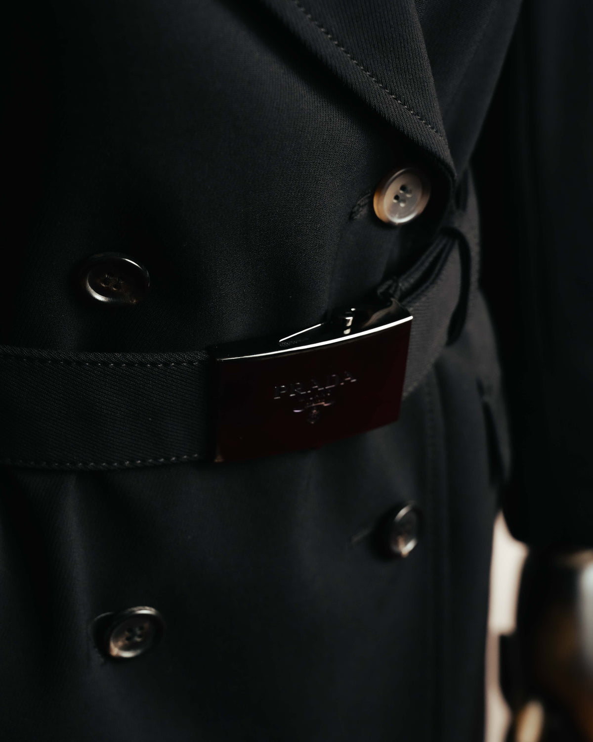 共布ベルトに配された、PRADA MILANO DAL 1913の文字と紋章が刻まれた黒系メタルの大型バックルを捉えたディテールカット。周囲にはダブルブレストのボタンとフラップポケットの縁が写ります。