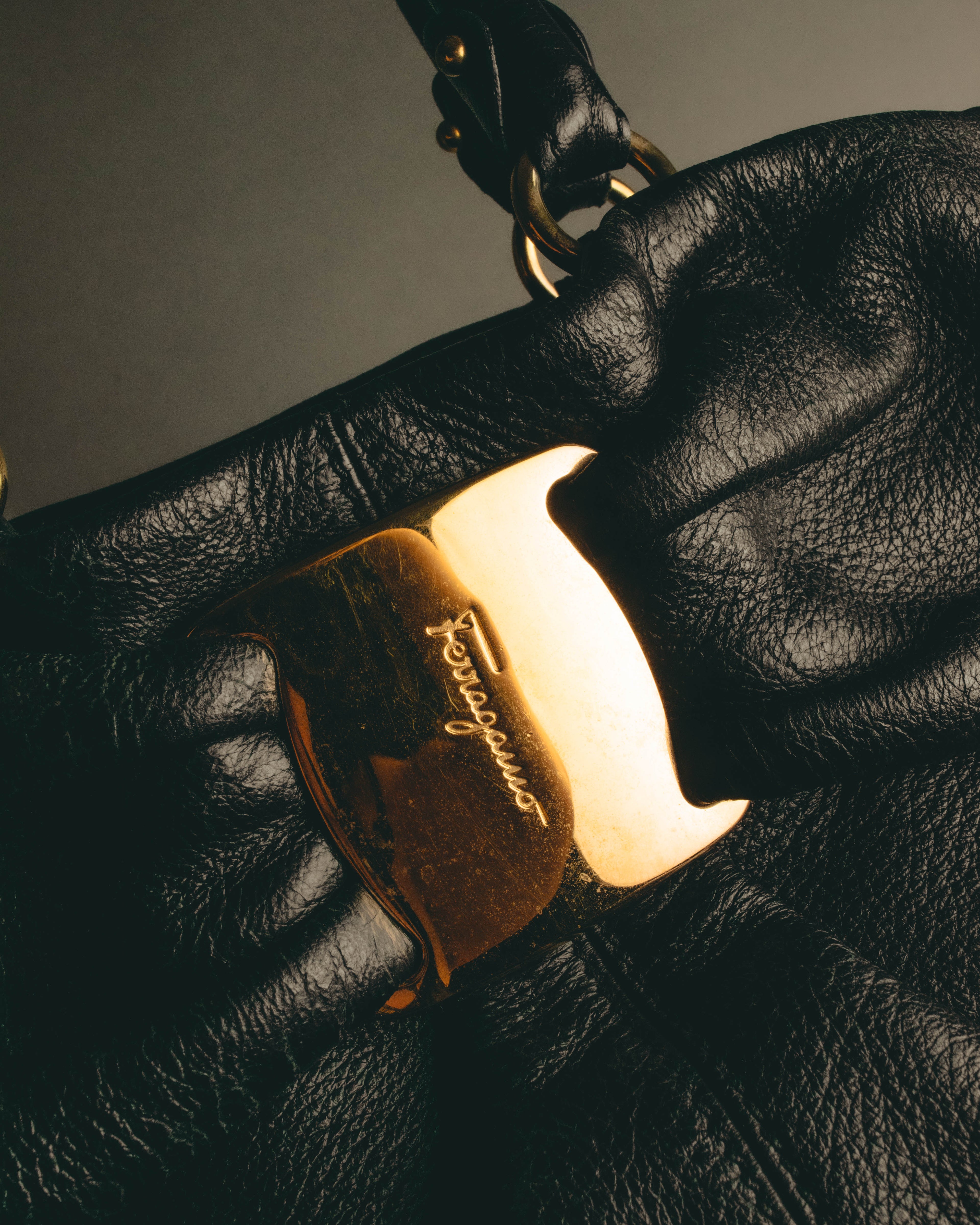 中央に筆記体のFerragamoロゴを刻んだ大型ゴールドトーンの留め具（クラスプ）と、丸環を用いたトップハンドル付け根を捉えたディテールカット。細やかなシボが浮かぶブラックレザーと深いフロントギャザーも確認できます。