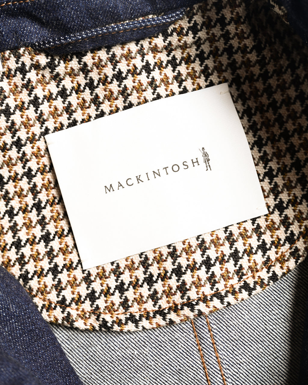 MACKINTOSH表記のブランドタグと、アイボリー・ブラックを基調にブラウンを交えた襟裏の千鳥格子調チェックを写したカット。周囲には濃紺デニムと橙からブラウン系のステッチも確認できます。