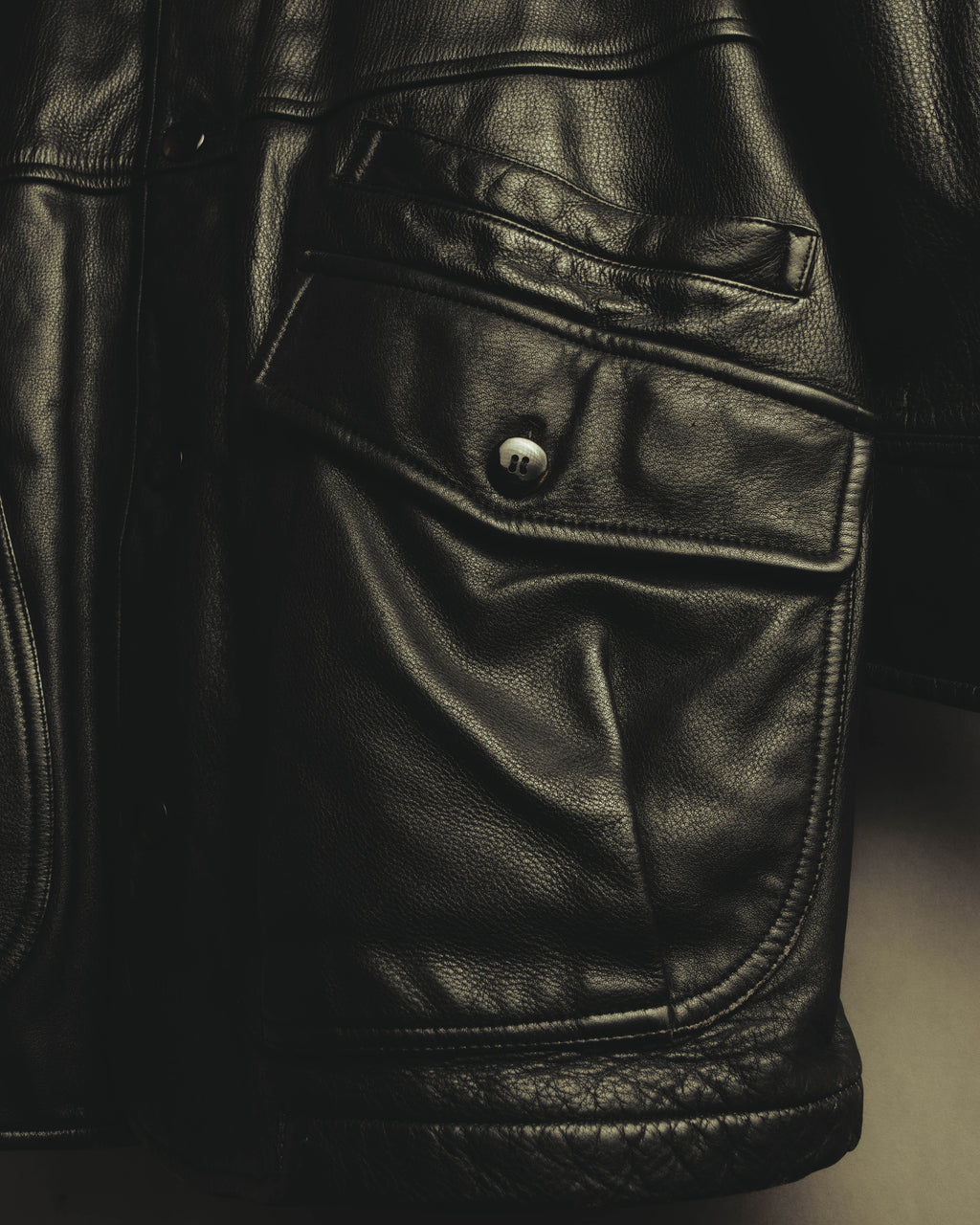 ブラックレザーの粒状のシボと、両腰に配された立体的なフラップポケットを捉えたディテールカット。フラップは4穴ボタンで留める構造です。