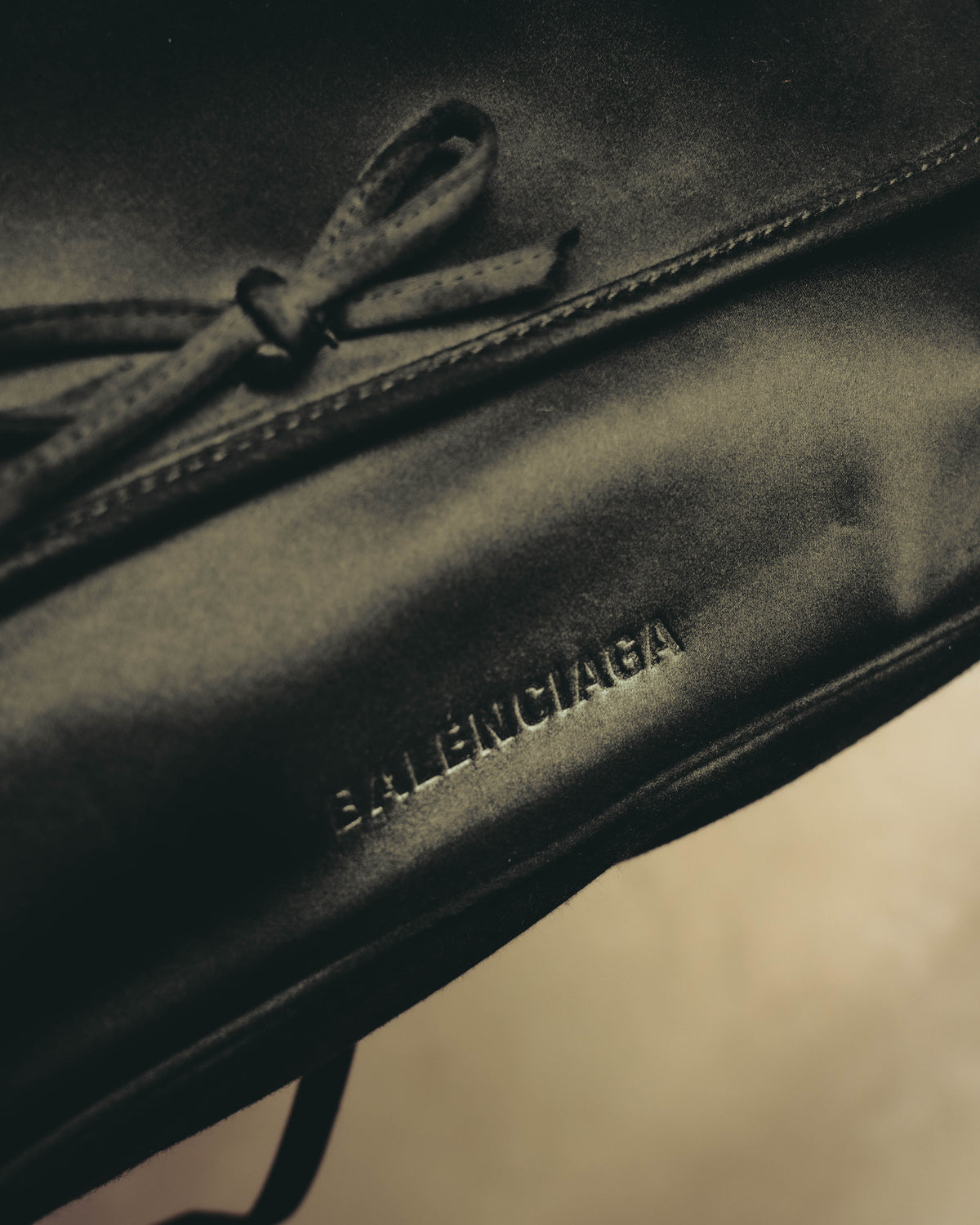 同色のリボンを配したフラップ中央から、前面下部のBALENCIAGA型押しロゴまでを捉えたディテールカット。サテン系素材の繊細な表情と縁のステッチも確認できます。
