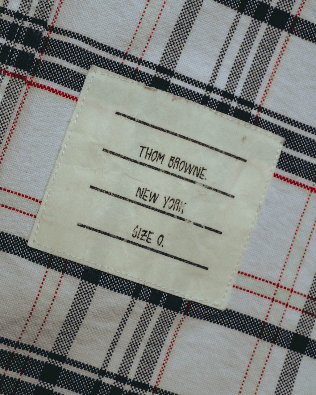タータンチェックのフロント裾に配された外付けブランドネームのアップ。「THOM BROWNE. NEW YORK.」「Size 0」の表記が確認できる。
