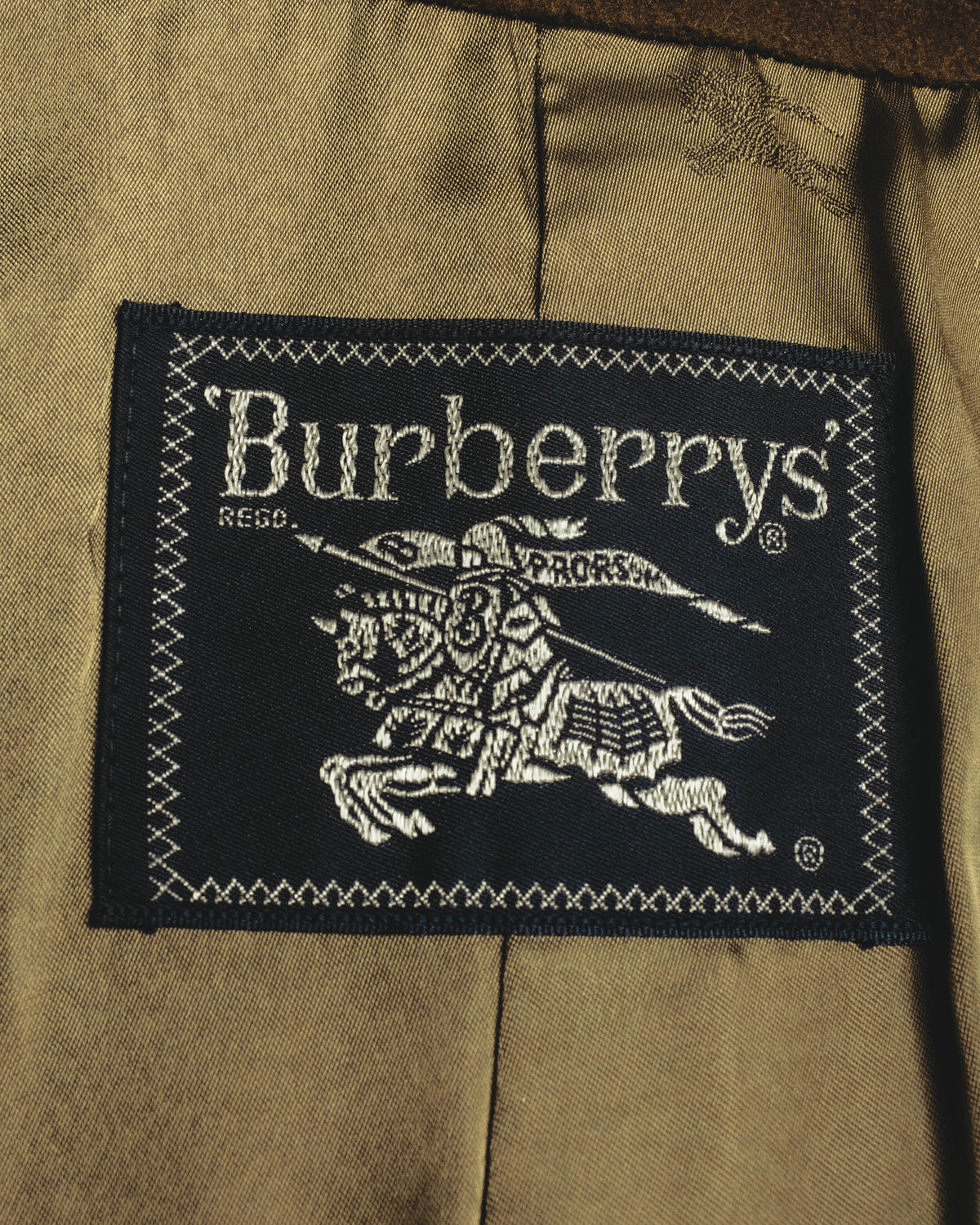 内側に配された、黒地に旧表記のBurberrys'と騎馬騎士図案を織り込んだ主タグのクローズアップ。キュプラ裏地の上に縫い付けられています。