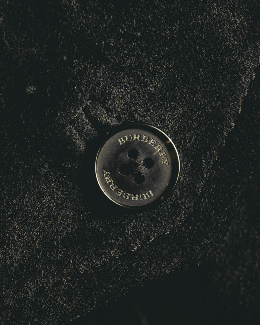 スエード調の起毛感を備えた表地に配された、BURBERRY刻印入りの4つ穴ボタンのアップ。毛並みによる濃淡とボタンの刻印を確認できるディテールカット。