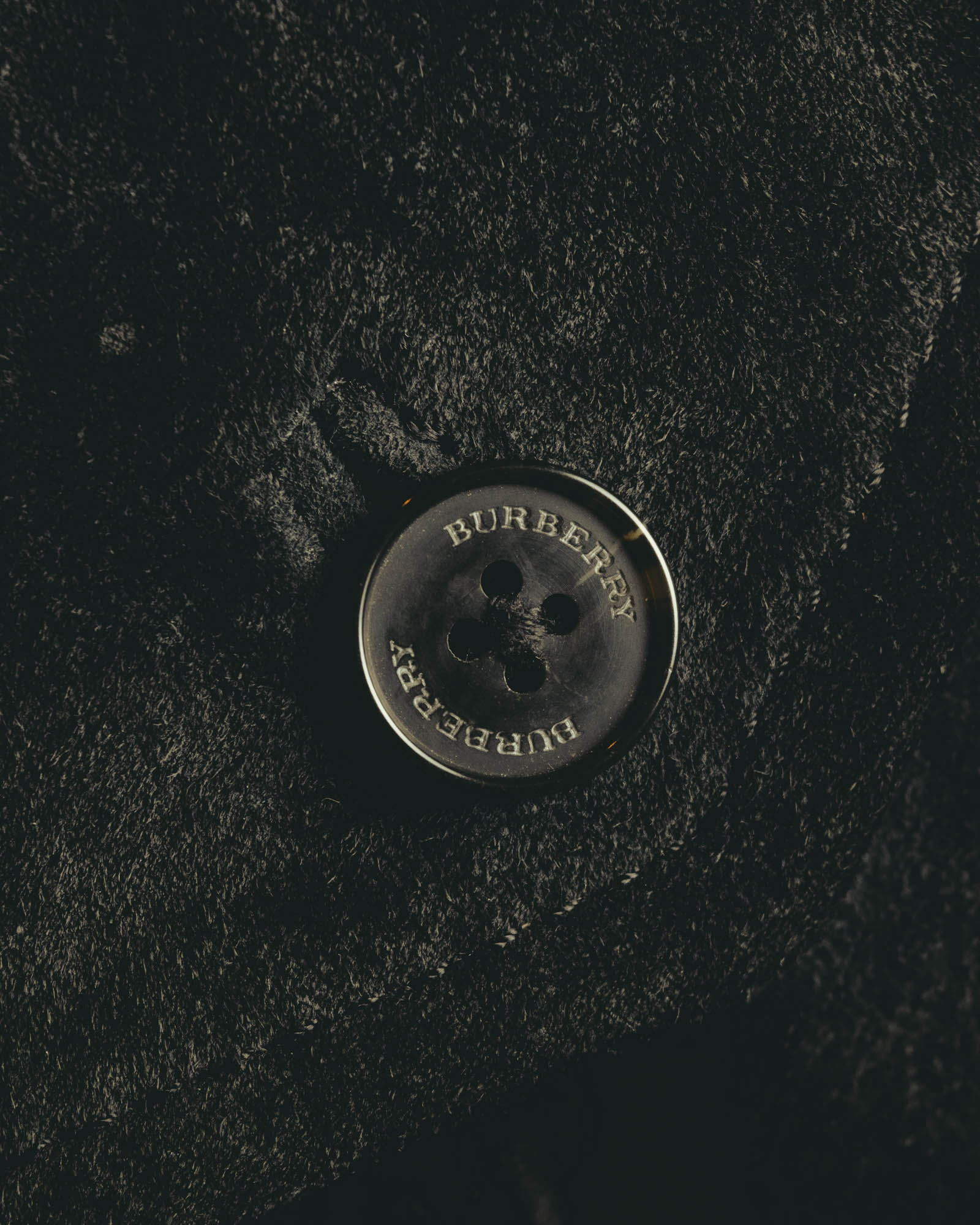 スエード調の起毛感を備えた表地に配された、BURBERRY刻印入りの4つ穴ボタンのアップ。毛並みによる濃淡とボタンの刻印を確認できるディテールカット。
