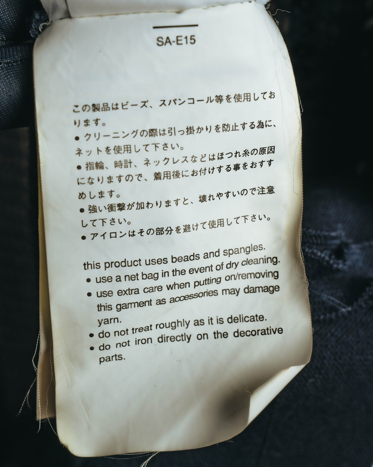 品番「SA-E15」と、ビーズ・スパンコール使用製品のケア方法を記した注意書きタグ。装飾部分へのアイロンを避ける旨とクリーニング時の注意が日英で確認できます。