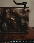 深いカカオブラウンの外装に施された、同心状の凹凸が連なる亀甲状エンボスのディテール。上部にはショルダーストラップと本体をつなぐダークカラーのリングパーツを確認できます。