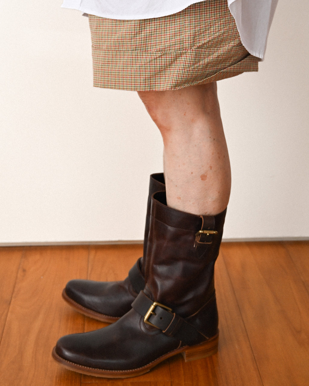 ショートパンツにダークブラウンのプレントゥ エンジニア レザーブーツを合わせた足元の着用カット。甲部分と筒上部に配された真鍮製バックルベルトが確認できます。