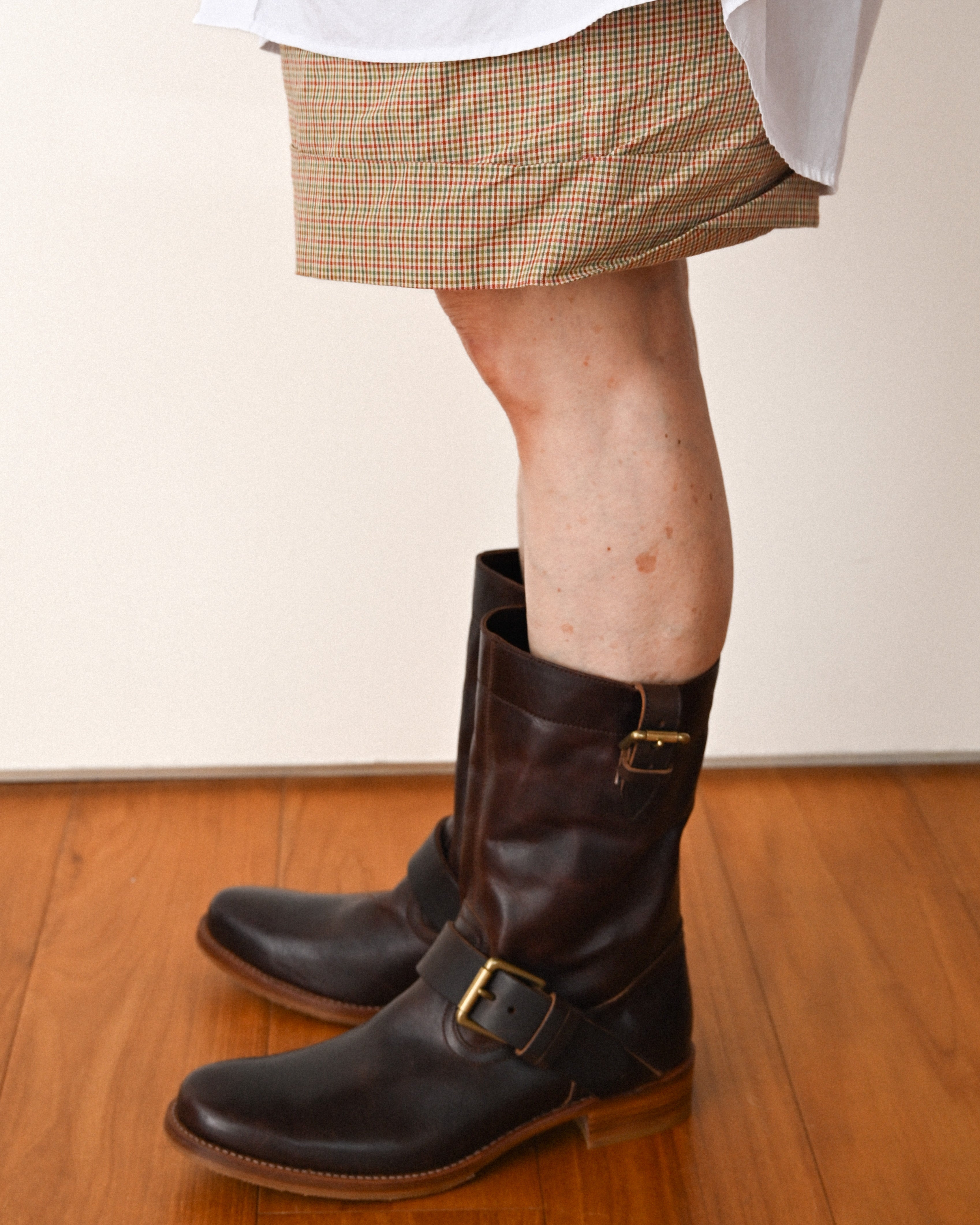 ショートパンツにダークブラウンのプレントゥ エンジニア レザーブーツを合わせた足元の着用カット。甲部分と筒上部に配された真鍮製バックルベルトが確認できます。