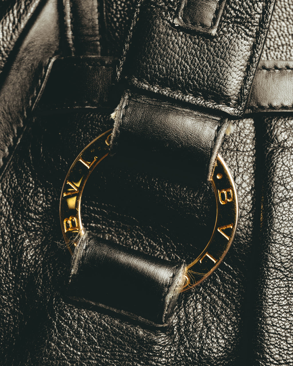 ダブルハンドルの付け根にあしらわれた、BVLGARIの刻印を反復したゴールドカラーのリング金具のディテールカット。ブラックのシボ状レザーと金具のコントラストが確認できます。