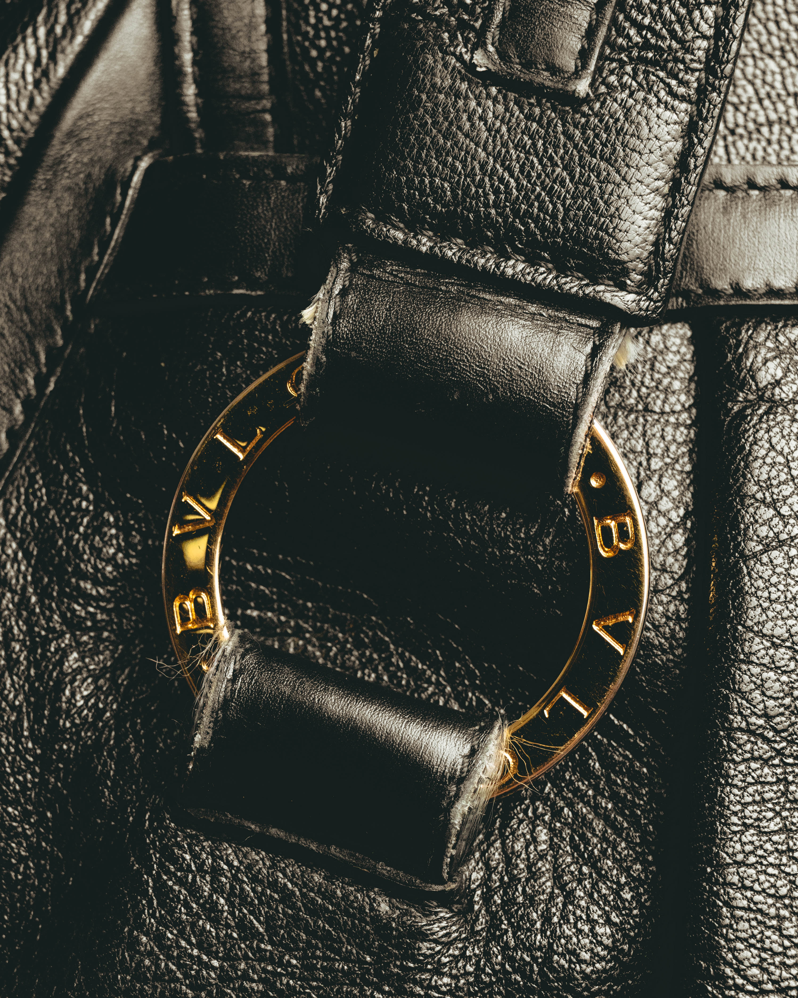 ダブルハンドルの付け根にあしらわれた、BVLGARIの刻印を反復したゴールドカラーのリング金具のディテールカット。ブラックのシボ状レザーと金具のコントラストが確認できます。