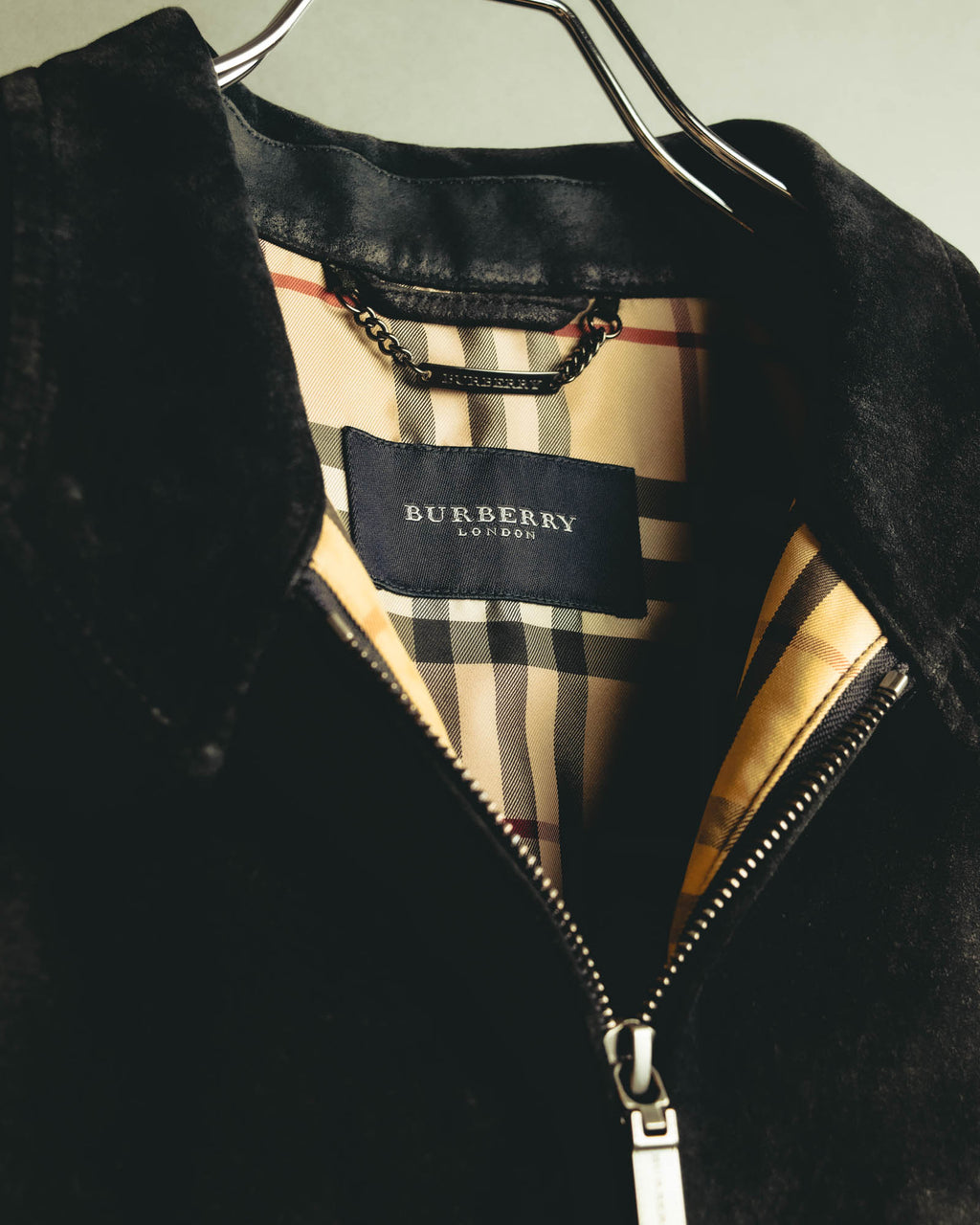 襟裏に配されたブランド刻印入りの金属製吊りチェーンプレートと、BURBERRY LONDONの織りタグを捉えたカット。ベージュ基調のチェック柄裏地とフロントジッパーも確認できる。