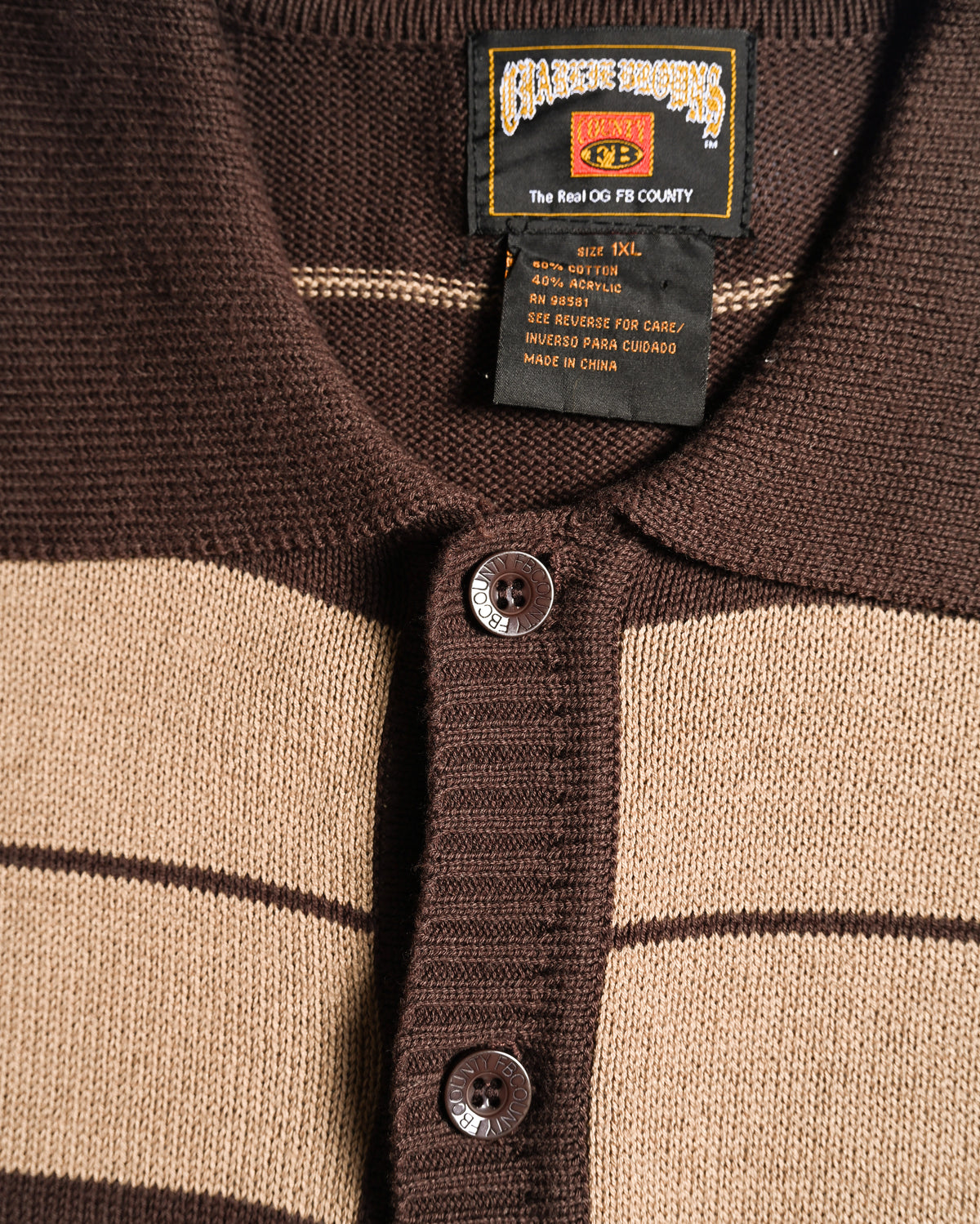 襟元のネックラベルと、サイズ1XL・綿60％／アクリル40％表記の品質表示タグを捉えたカット。フルボタン仕様の前立てにはFB COUNTY刻印入りボタンを配しています。
