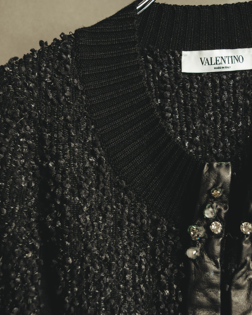 襟元のVALENTINO MADE IN ITALYネームと、ループ状の凹凸を持つブラックの編地を捉えたディテールカット。前立てには柔らかなブラックレザーと、爪留め状のクリアストーンが確認できます。
