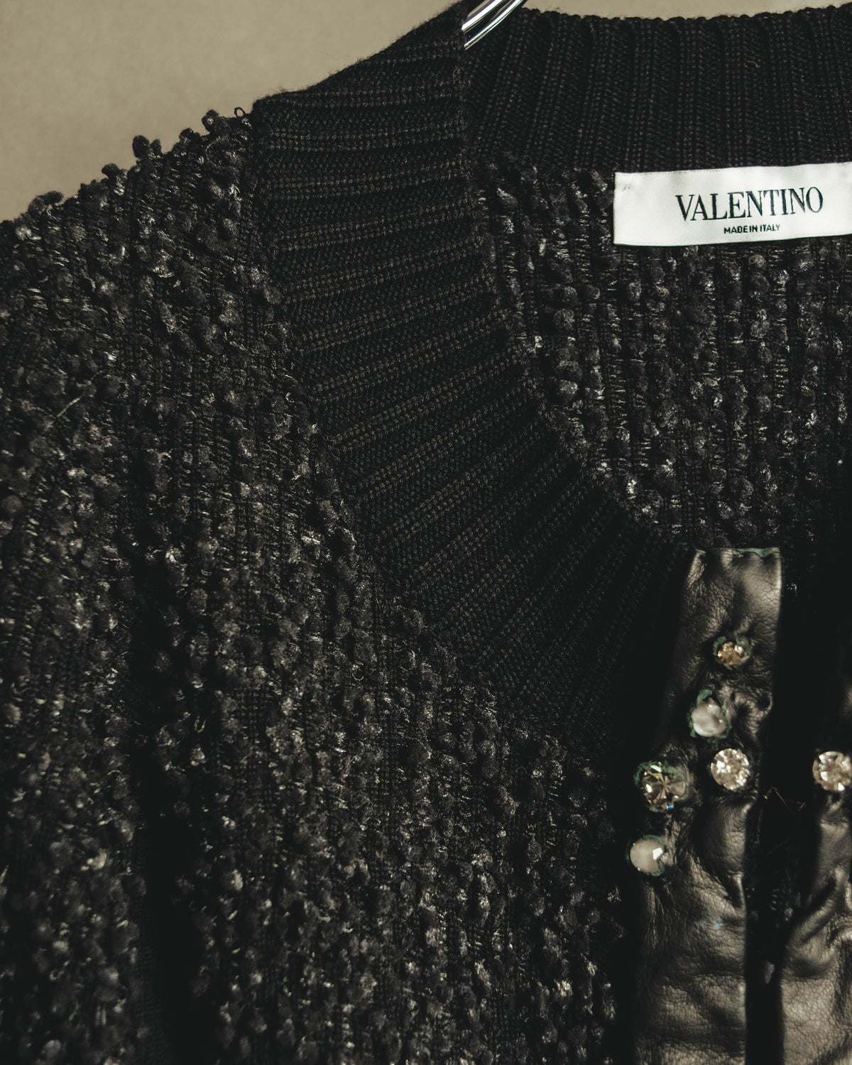 襟元のVALENTINO MADE IN ITALYネームと、ループ状の凹凸を持つブラックの編地を捉えたディテールカット。前立てには柔らかなブラックレザーと、爪留め状のクリアストーンが確認できます。