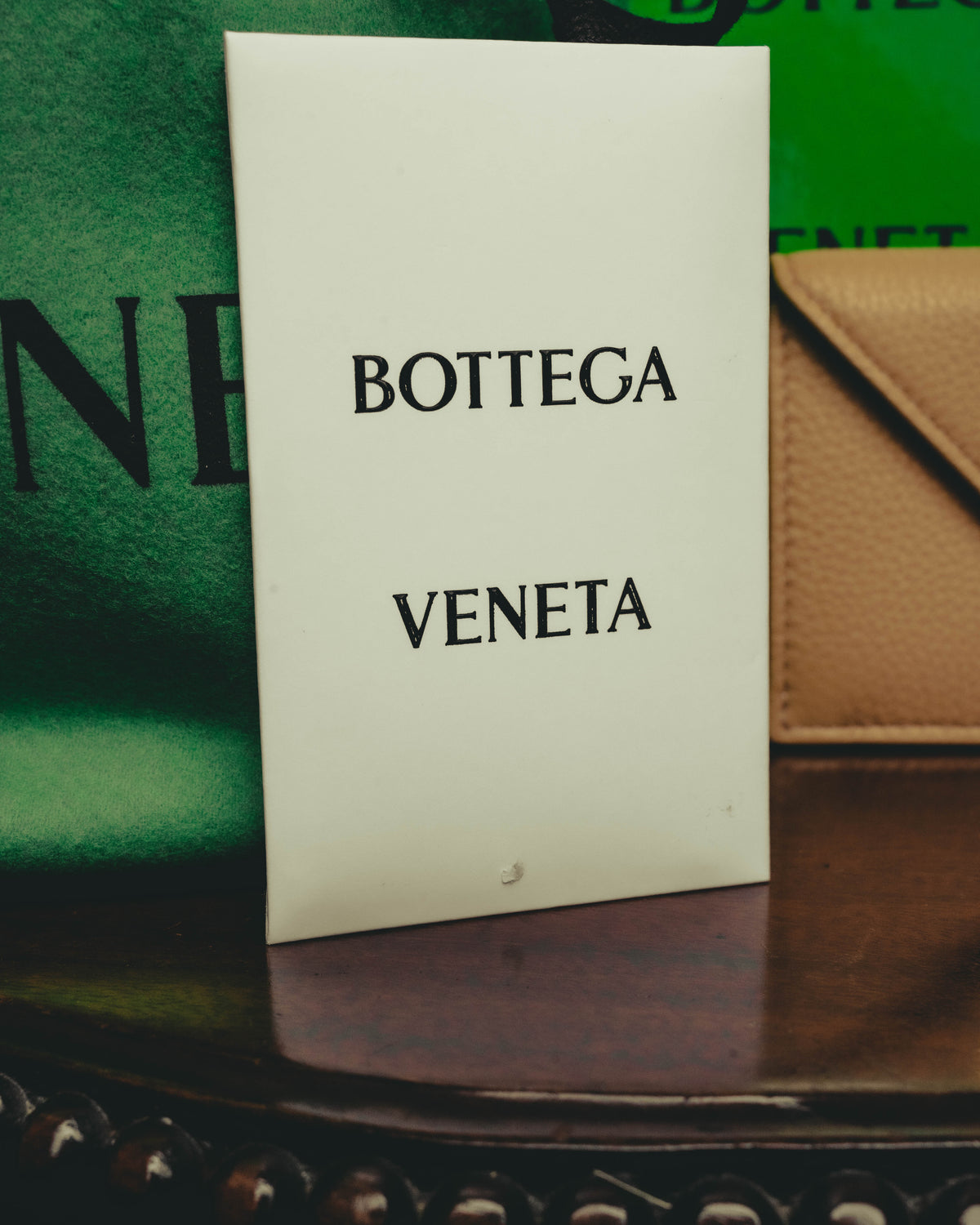 「BOTTEGA VENETA」のロゴが印字された白い付属ボックスを写したカット。背後にはグリーンカラーの付属品と、ベージュレザーの一部が確認できます。