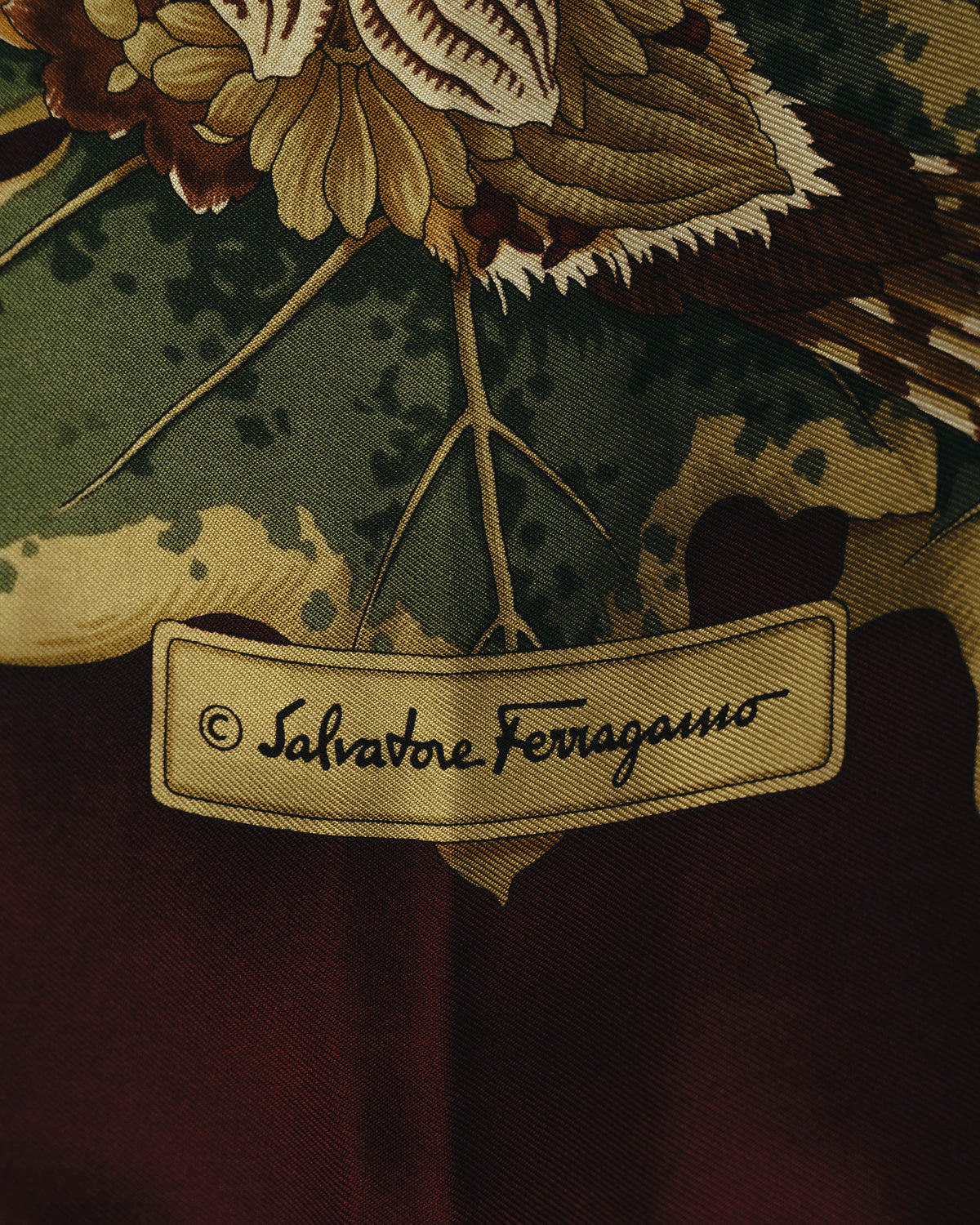 深いボルドーの地に、筆記体の「Salvatore Ferragamo」ロゴが配されたプリント部分のディテールカット。周囲には鳥の羽根や植物モチーフ、シルクの緻密な綾織りが確認できます。