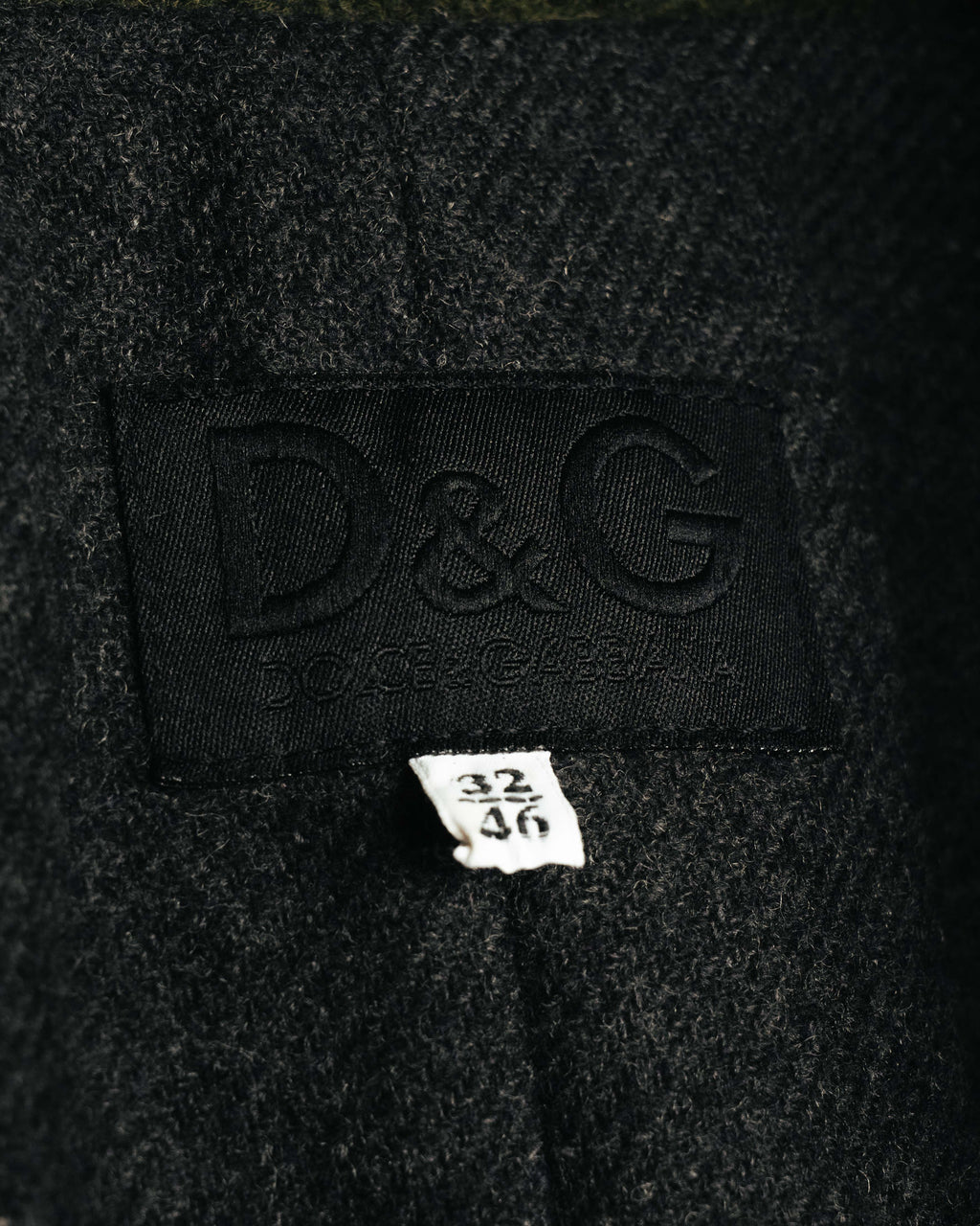 襟元に配されたD&G DOLCE & GABBANAのブランドタグと、32／46表記のサイズタグを捉えたカット。周囲には繊細な起毛感を伴うダークチャコールのウール混生地が確認できます。