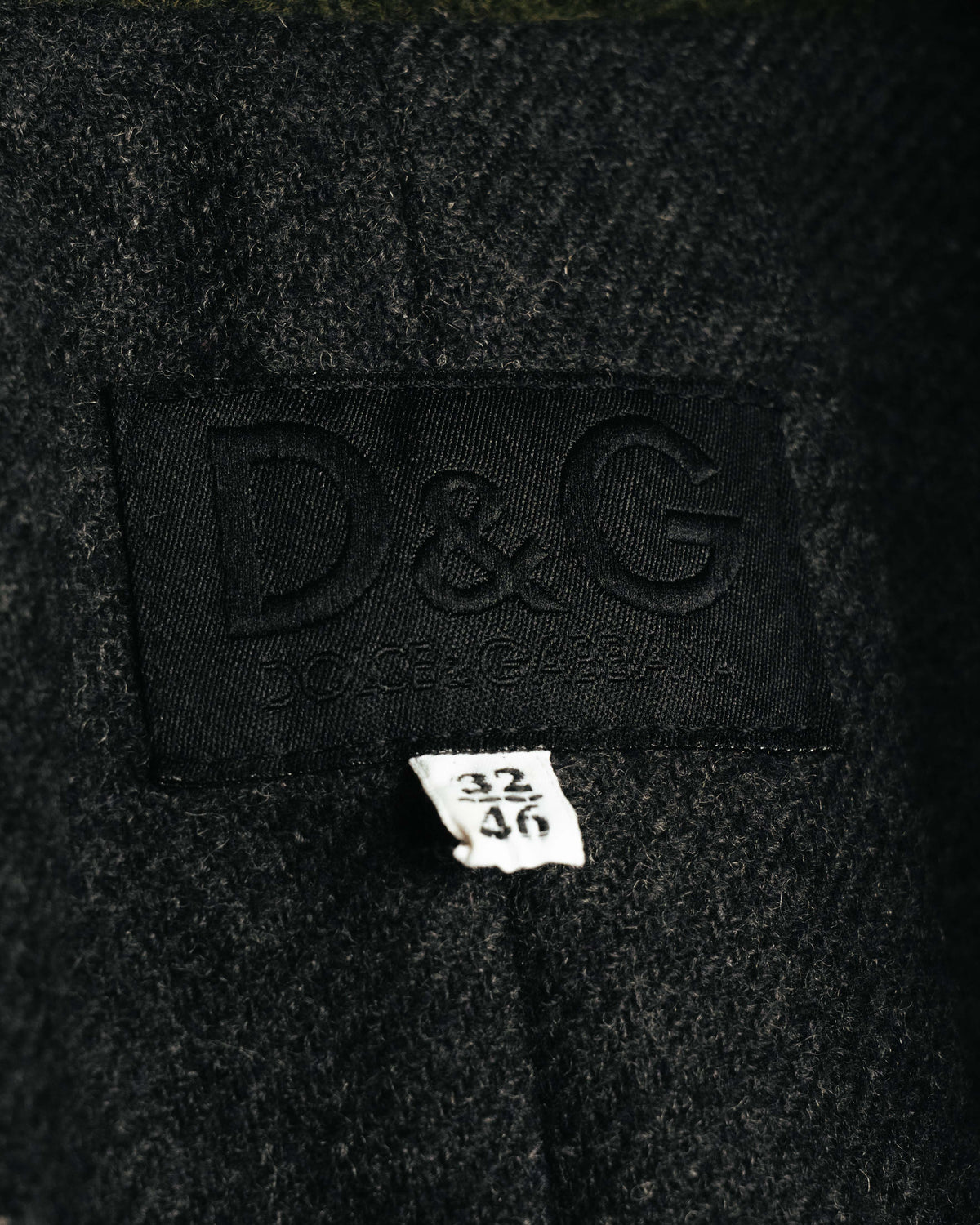 襟元に配されたD&G DOLCE & GABBANAのブランドタグと、32／46表記のサイズタグを捉えたカット。周囲には繊細な起毛感を伴うダークチャコールのウール混生地が確認できます。
