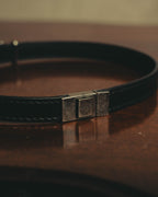 SAINT LAURENT PARISのロゴ刻印を配した、スクエア型メタル留め具（クラスプ）のディテール。ブラックレザーストラップと、ヴィンテージライクな風合いのシルバーカラー金具を確認できるカット。