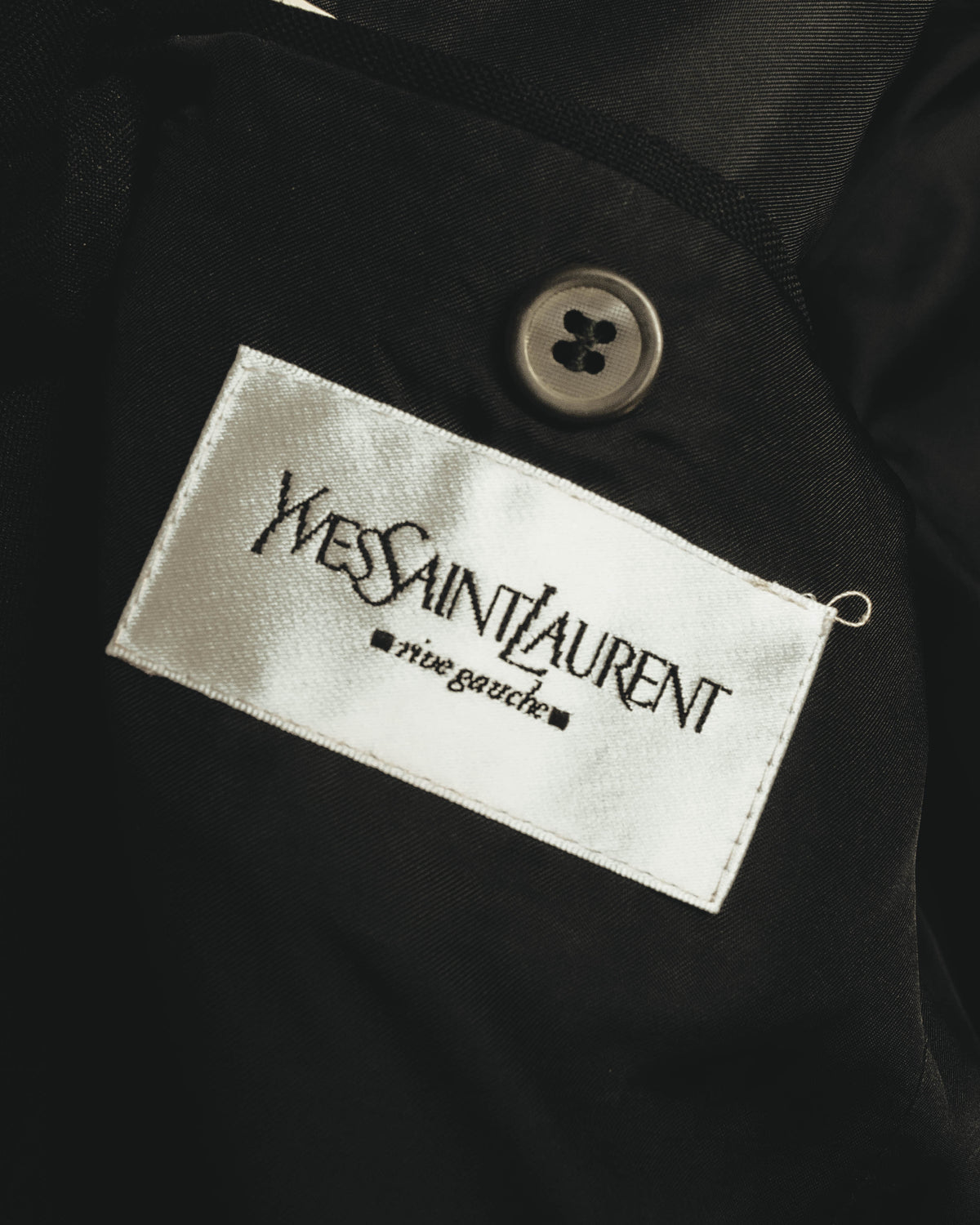 内側に配された白いサテン織りの「Yves Saint Laurent rive gauche」タグを捉えたカット。タグ上部には4つ穴のボタンとブラックの裏地が見える。