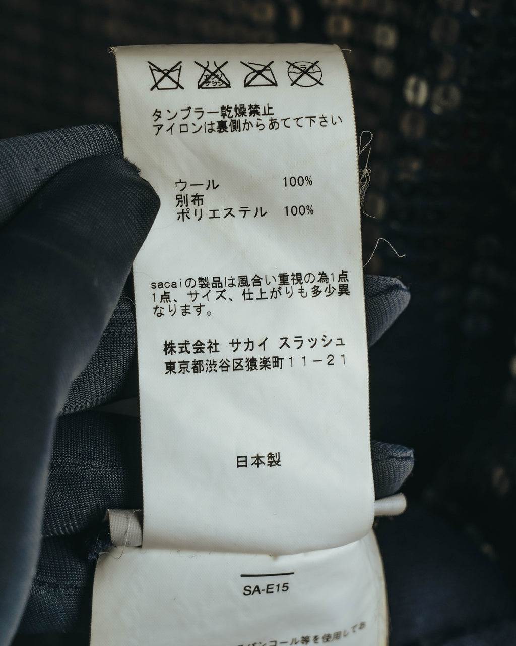 ウール100パーセント、別布ポリエステル100パーセントの素材表記を確認できる品質表示タグ。株式会社サカイスラッシュの住所表記と品番「SA-E15」が写るカット。
