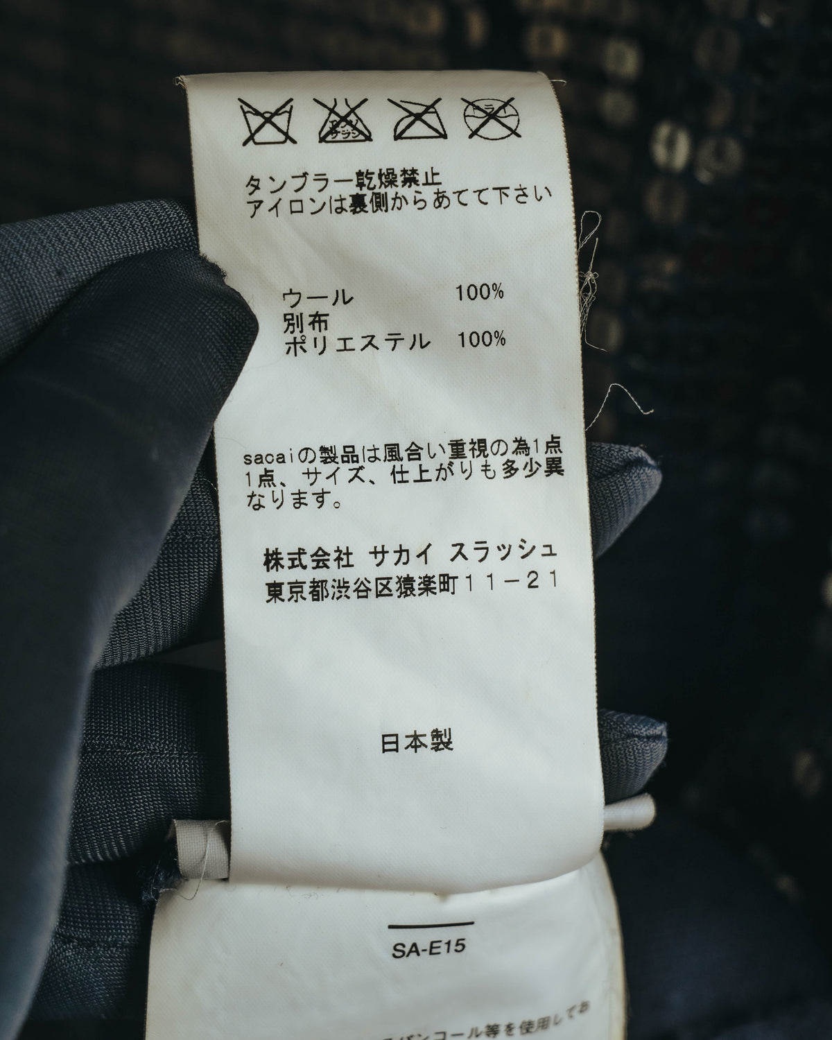 ウール100パーセント、別布ポリエステル100パーセントの素材表記を確認できる品質表示タグ。株式会社サカイスラッシュの住所表記と品番「SA-E15」が写るカット。