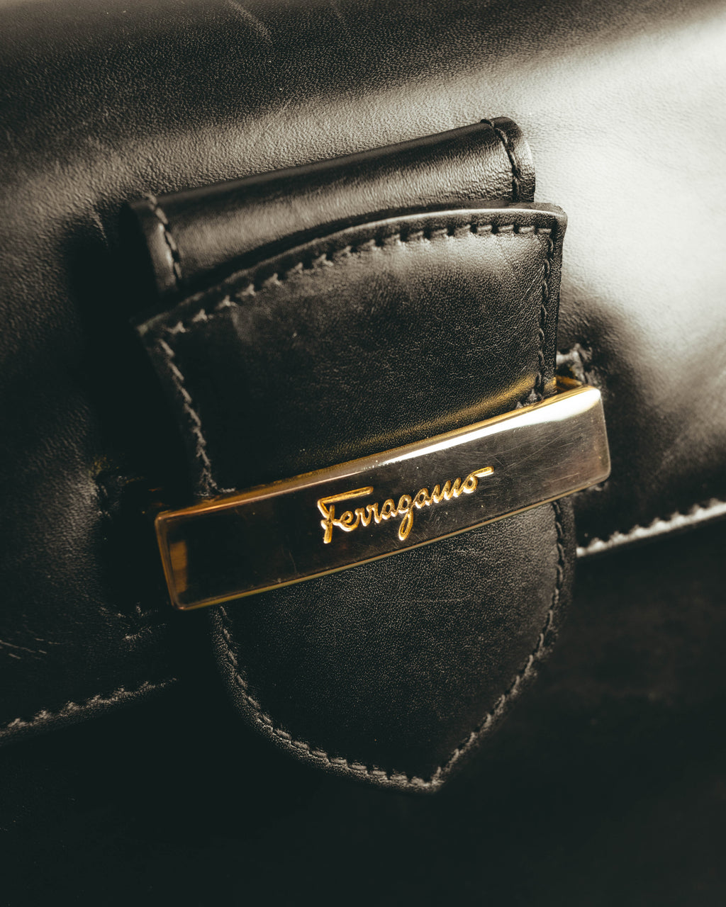 ブラックレザーのトップハンドル基部に配された、Ferragamoのスクリプトロゴ入りゴールドカラープレートのディテール。周囲のレザーパーツとステッチも確認できるカットです。