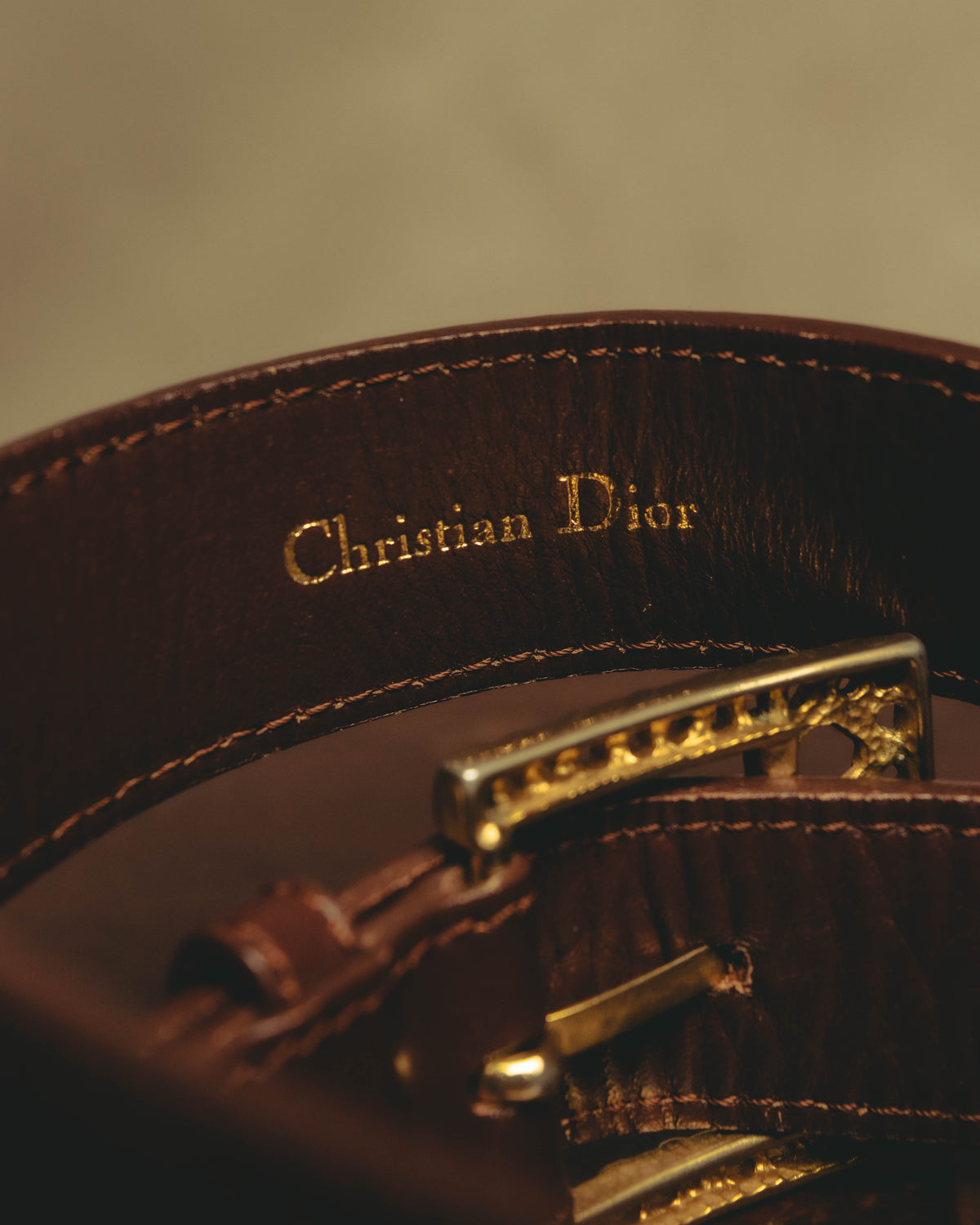 ダークブラウンの帯材内側に施された、セリフ体の「Christian Dior」金色箔押し表記を捉えたカット。端正なステッチと、手前にピン式バックルの裏側が写ります。