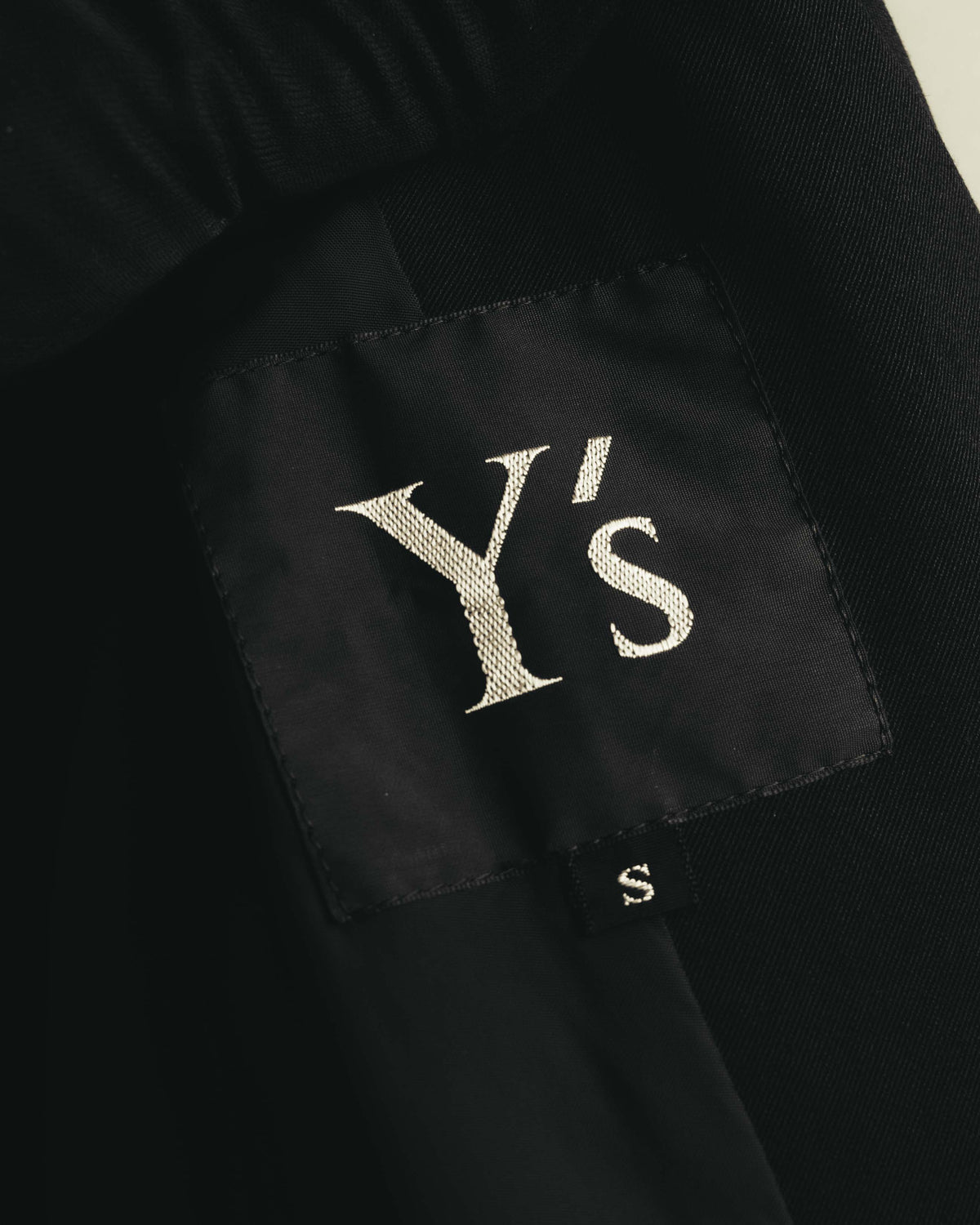黒地に白糸のセリフ体で織られた「Y's」ブランドネームと、別体のアルファベットサイズタグのクローズアップ。サイズ表記はS。