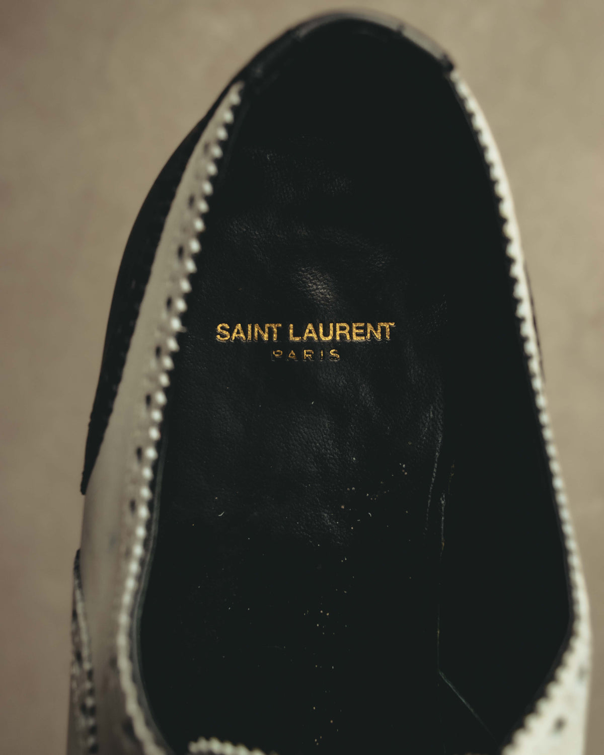 ブラックのインソール中央に入る、ゴールドカラーの「SAINT LAURENT PARIS」ロゴ刻印を写したディテールカット。履き口周囲にはホワイトレザーとブローギングのパーフォレーションが確認できます。