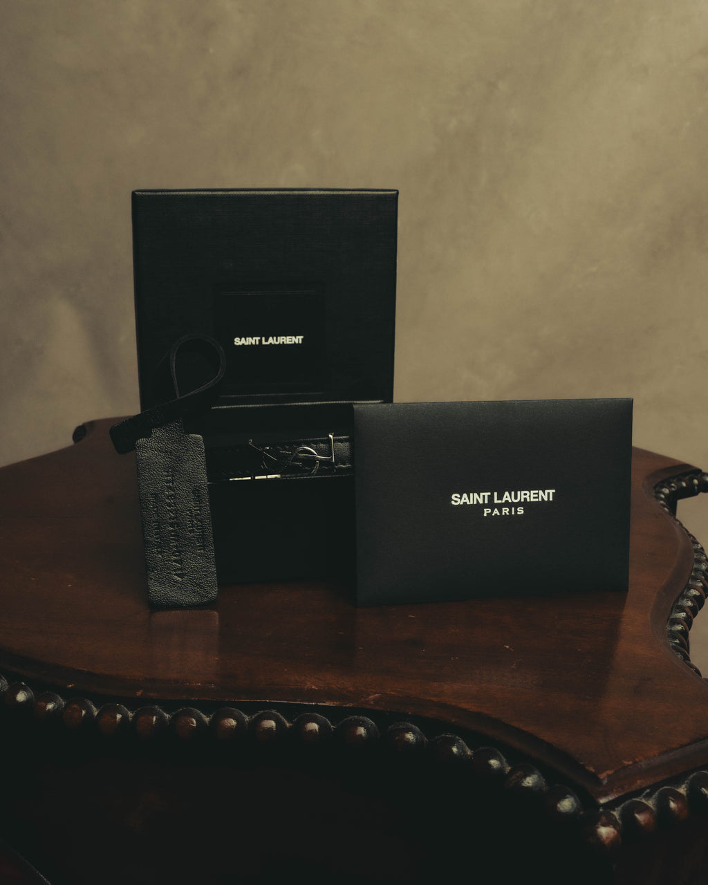 ブラックレザーのダブルラップブレスレットを、SAINT LAURENT PARISロゴ入りの付属ボックスおよび保存袋とともに写した付属品を含む全体カット。中央にはシルバーカラーのYSLモノグラムが確認できます。