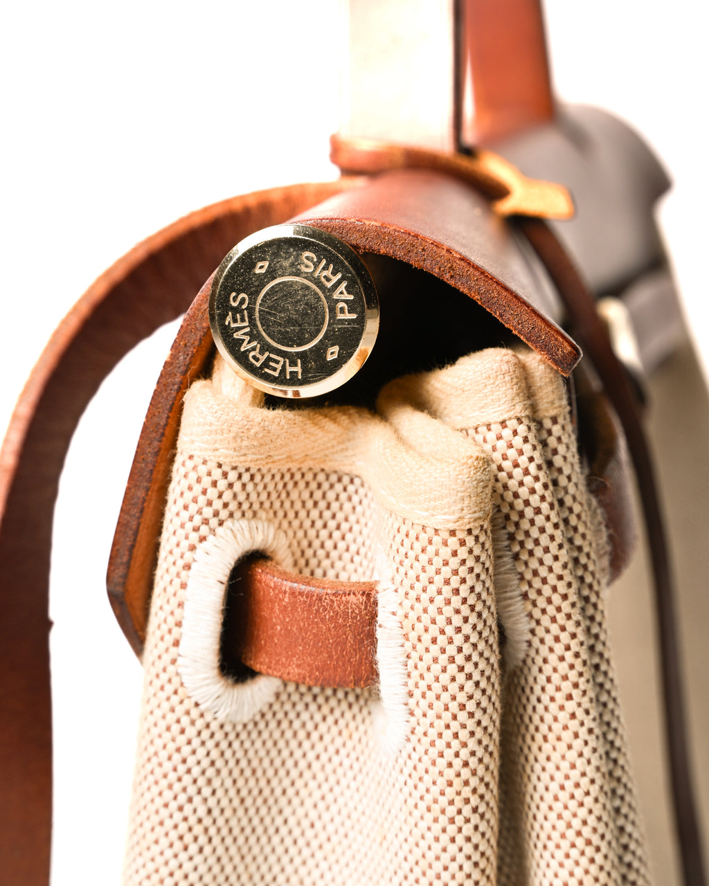 フロントのベルトを通して留める開閉構造を捉えた、HERMÈS PARIS刻印入りゴールドカラーのセリエ金具のアップ。生成りからライトベージュの織物製ボディと深いブラウンのレザーが交差する箇所です。