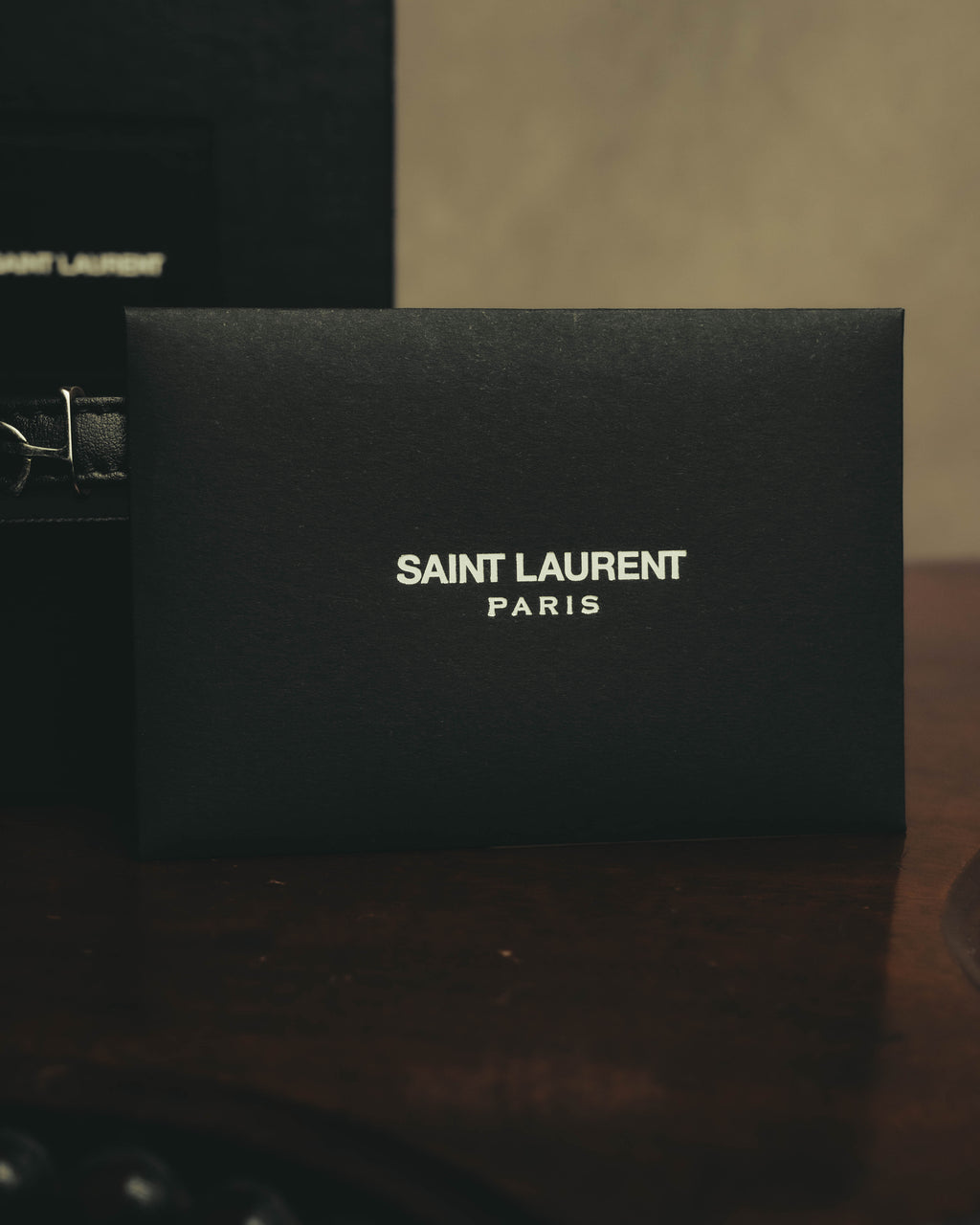 SAINT LAURENT PARISのロゴが配されたブラックの付属ボックスを写したカット。背後にはレザーストラップの一部と留め具（クラスプ）が確認できます。