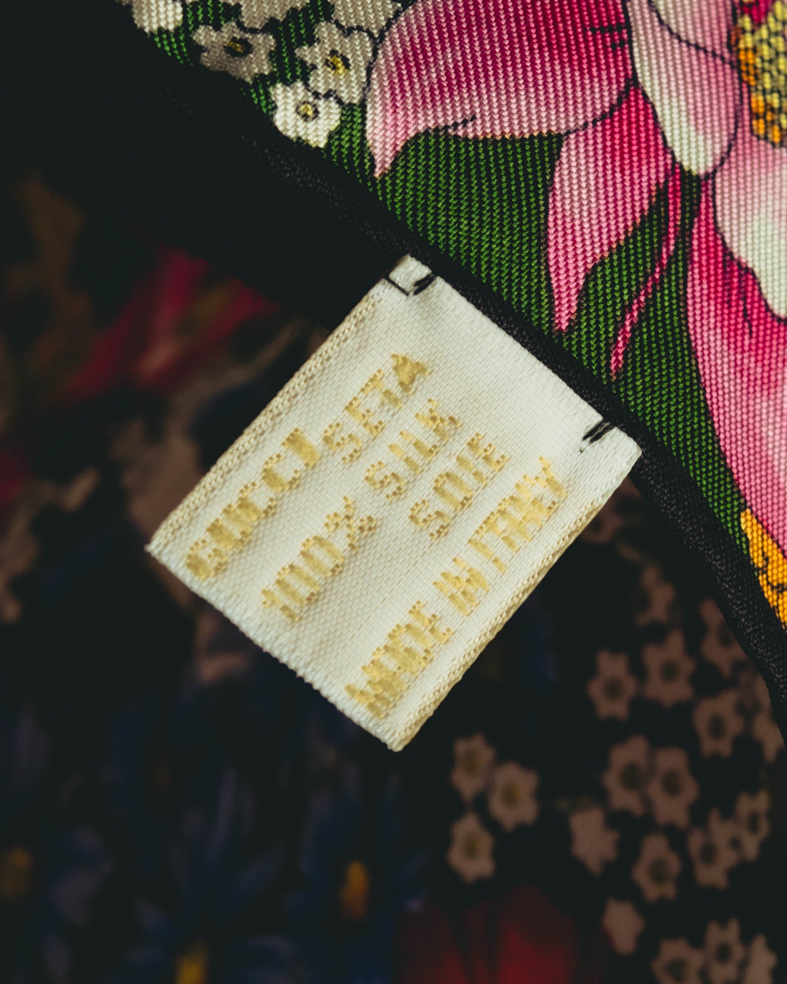 GUCCI、100% SILK、SOIE、MADE IN ITALYの表記が確認できる生成り色の織りタグ。ネイビー地に色彩豊かな花々が配された本体に縫い付けられています。