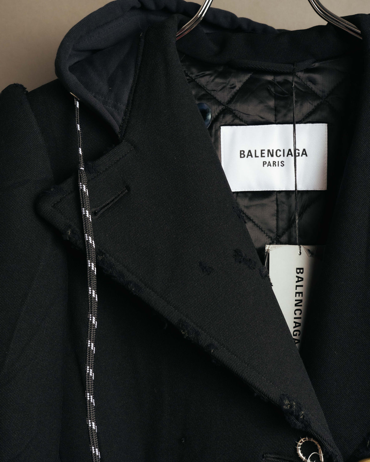 襟元に配された白地に黒文字の「BALENCIAGA PARIS」タグと、内側のキルティング仕立ての裏地を捉えたカット。チャコールブラック調のフードとフロント端の裁ち切り処理も確認できます。