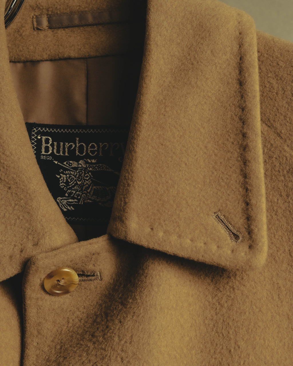 旧来のBurberrysロゴ、REGD.表記と騎士図案が確認できる内タグを、キャメルベージュの起毛した表地と首元のボタンとともに捉えたディテールカット。
