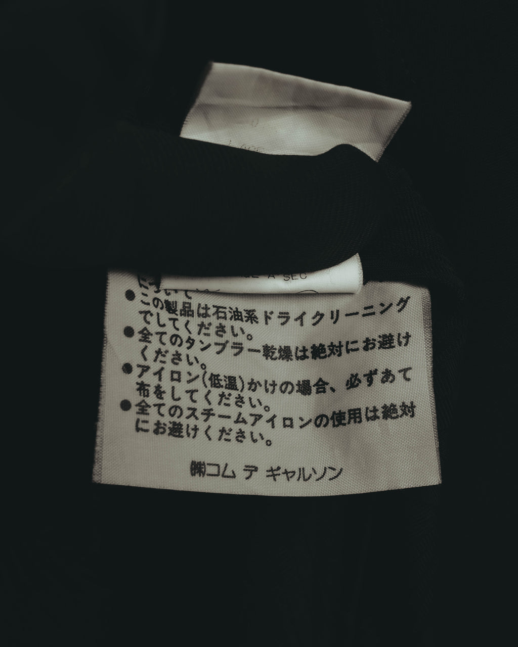 日本語でドライクリーニングやアイロン時の注意事項が記された内側の洗濯表示タグ。下部に「株式会社コム デ ギャルソン」の表記が確認できる。