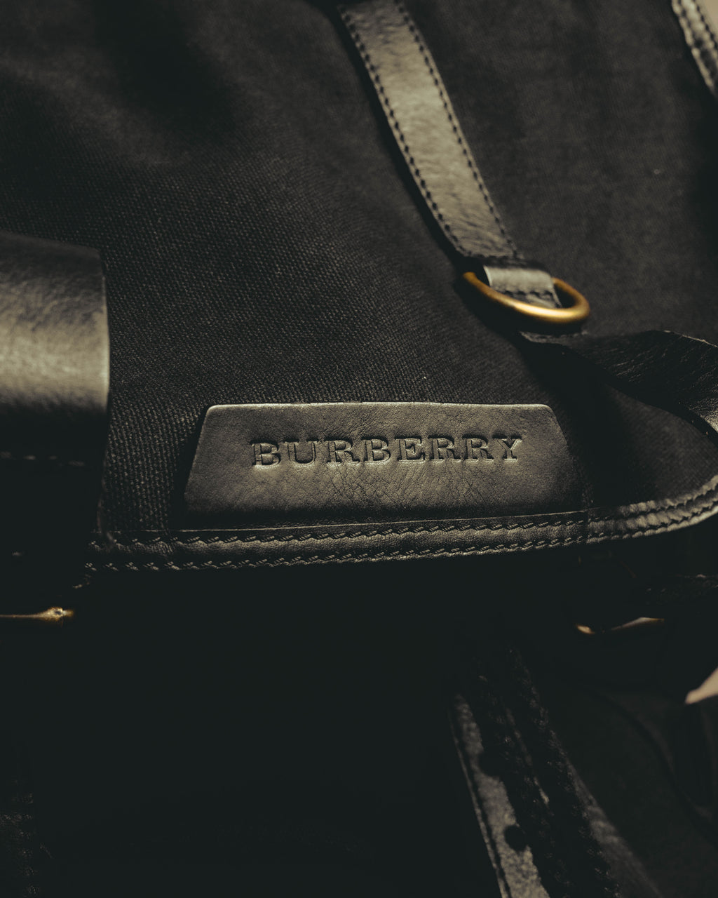 フラップ中央に配したBURBERRY刻印入りレザーパッチと、上部のDリングを捉えたディテールカット。本体の織り目とレザートリミングの質感差も確認できます。