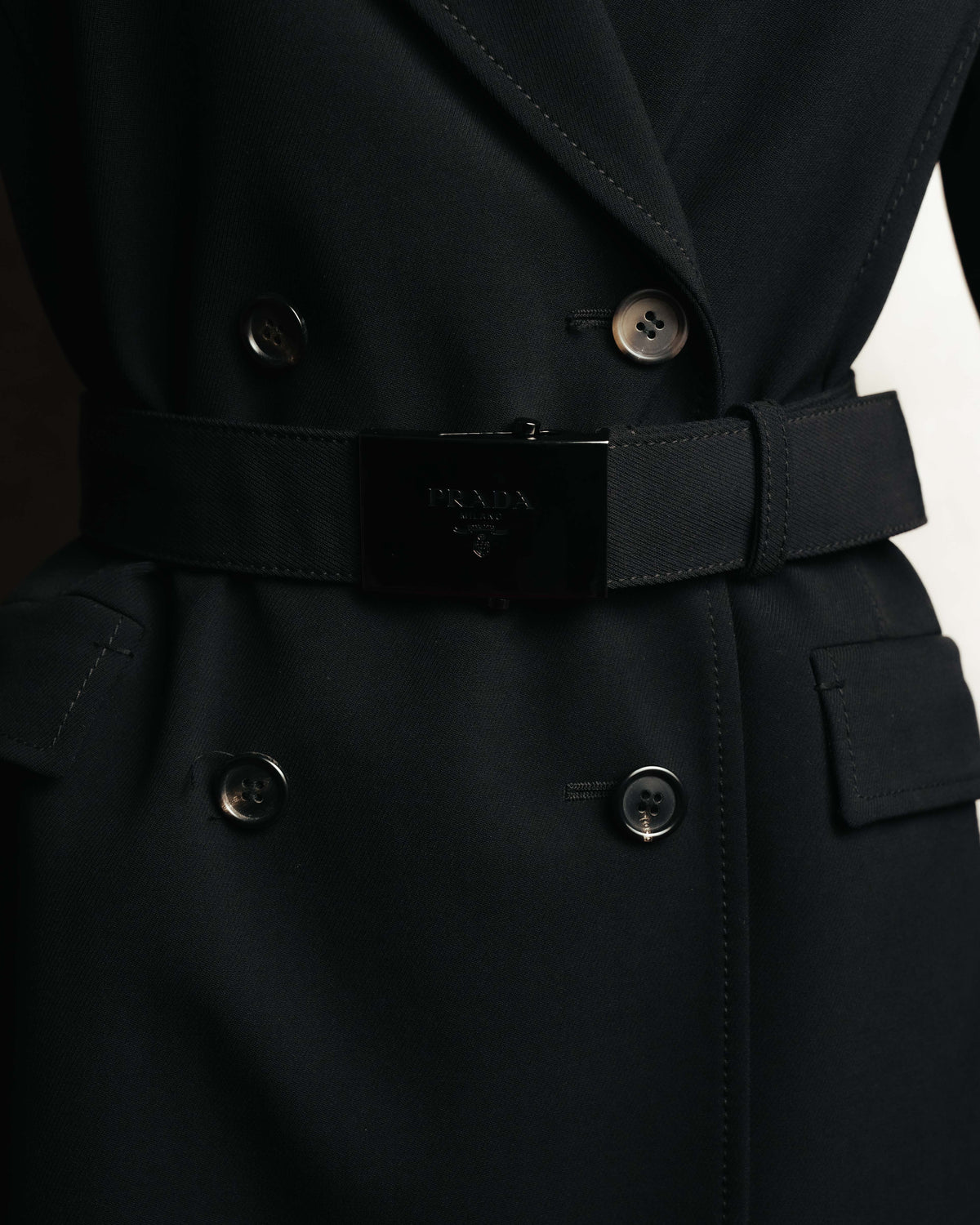 共布ベルトに配された、PRADA MILANO DAL 1913の文字と紋章が刻まれた艶を抑えた黒系メタルの大型バックル。ダブルブレストのボタンとフラップポケットも併せて確認できるディテールカット。