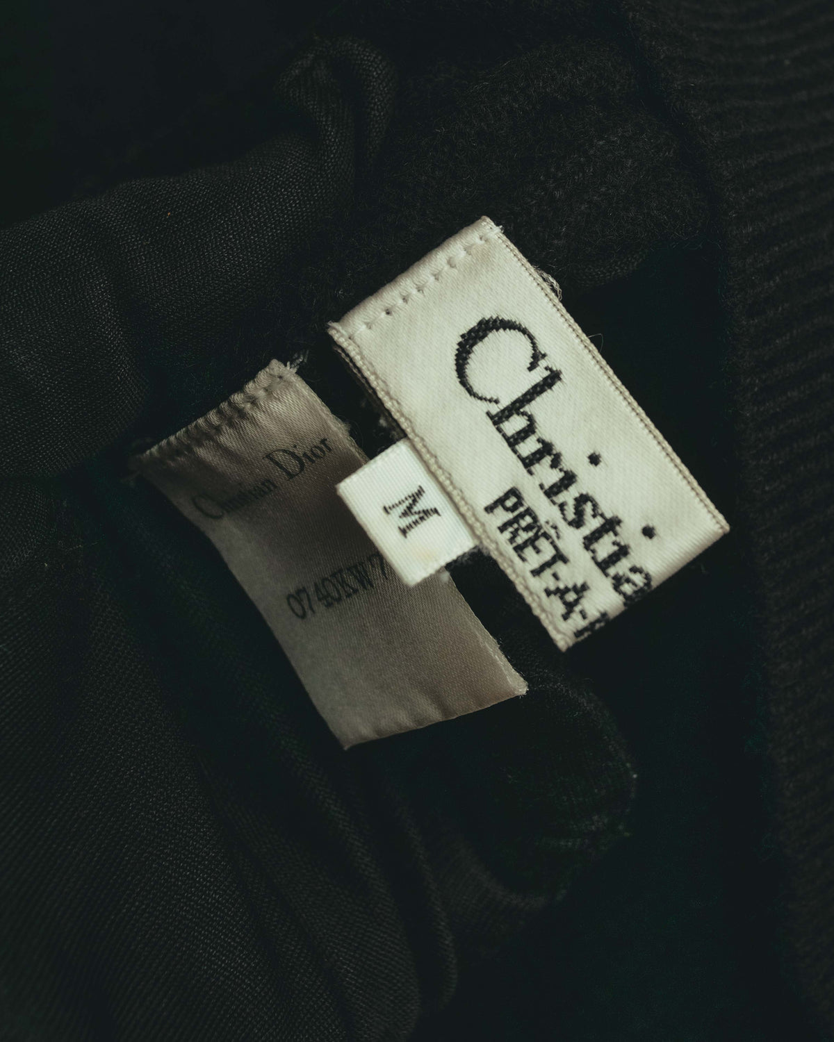 白地にChristian Dior PRET-A-PORTERと記された織りネームと、独立したMサイズ表記を捉えた内タグのカット。ブラックのニット地に取り付けられた副資材構成が確認できます。