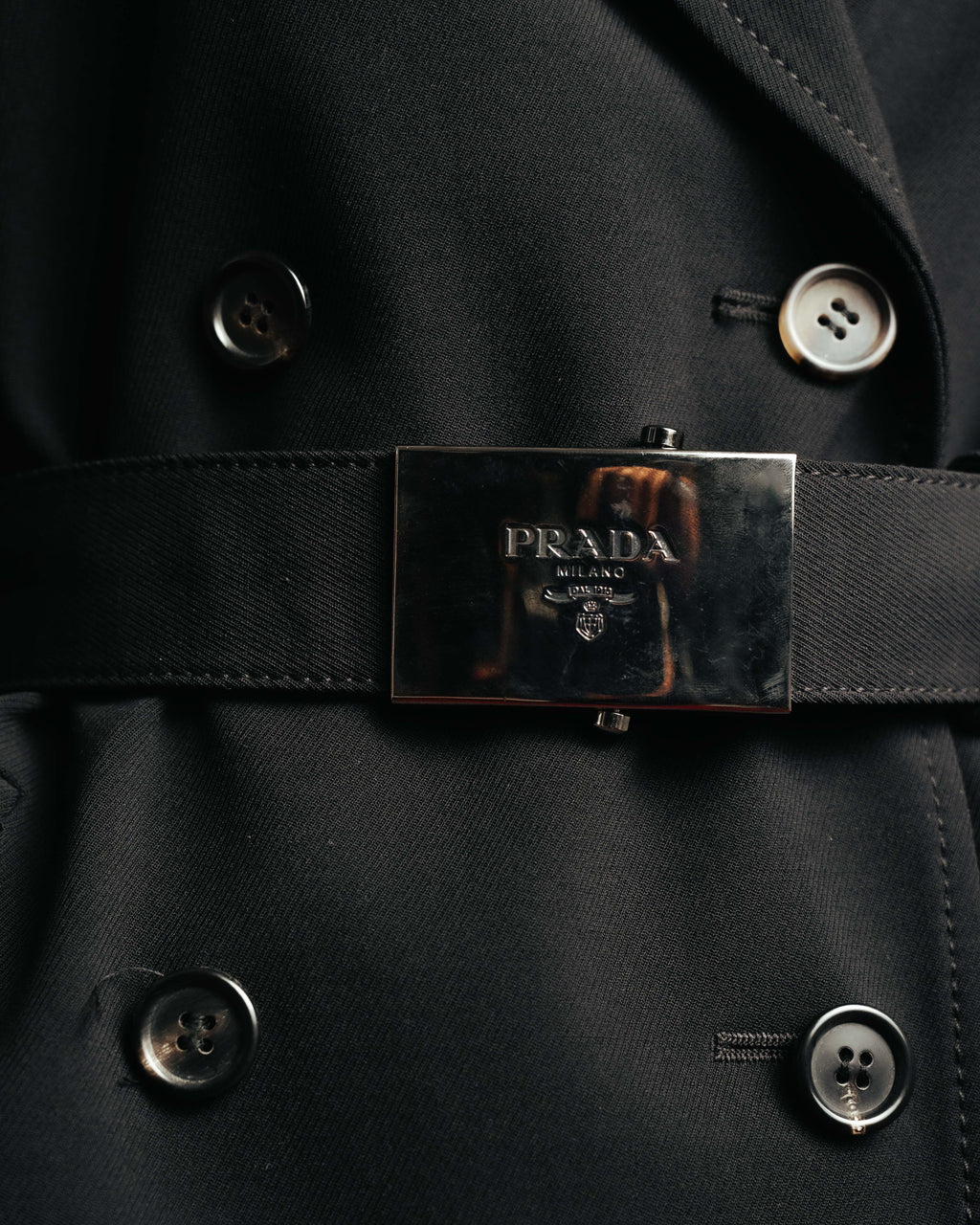 共布ベルトに配された、PRADA MILANO DAL 1913の文字と紋章を刻んだ黒系メタルの大型バックル。ダブルブレストのボタンとあわせて、フロントの構築的なディテールを捉えたカットです。