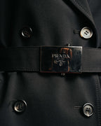 共布ベルトに配された、PRADA MILANO DAL 1913の文字と紋章を刻んだ黒系メタルの大型バックル。ダブルブレストのボタンとあわせて、フロントの構築的なディテールを捉えたカットです。