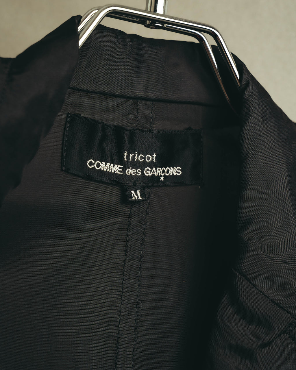 首元の内タグに「tricot COMME des GARÇONS」表記とサイズ「M」を確認できるカット。深いチャコールの滑らかな裏地と中央の縫製も写しています。