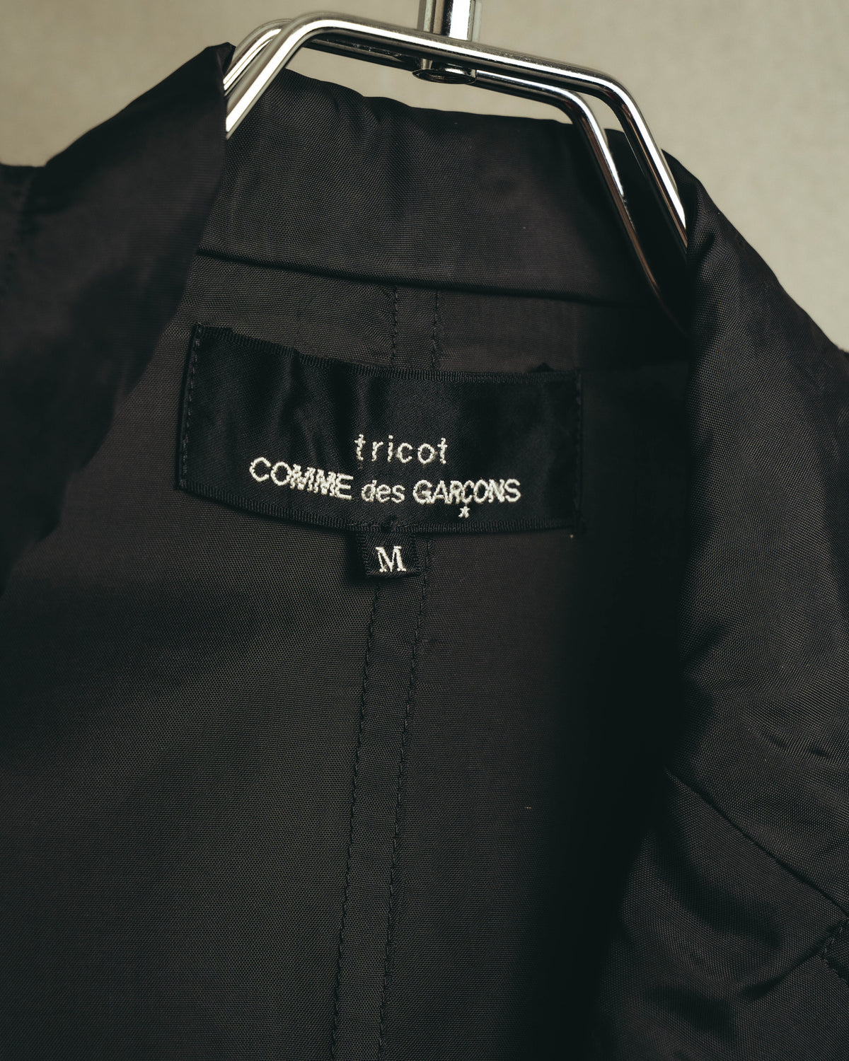 首元の内タグに「tricot COMME des GARÇONS」表記とサイズ「M」を確認できるカット。深いチャコールの滑らかな裏地と中央の縫製も写しています。
