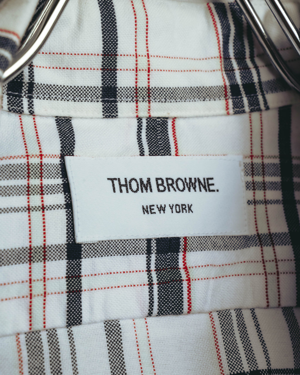 ホワイト、ブラック、グレーに細いレッドラインが交差するタータンチェック地に配された、「THOM BROWNE. NEW YORK」表記のネームタグ。